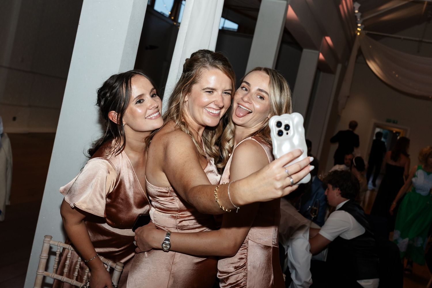 Three bridesmaids in champagne satin dresses take a selfie together at wedding reception.