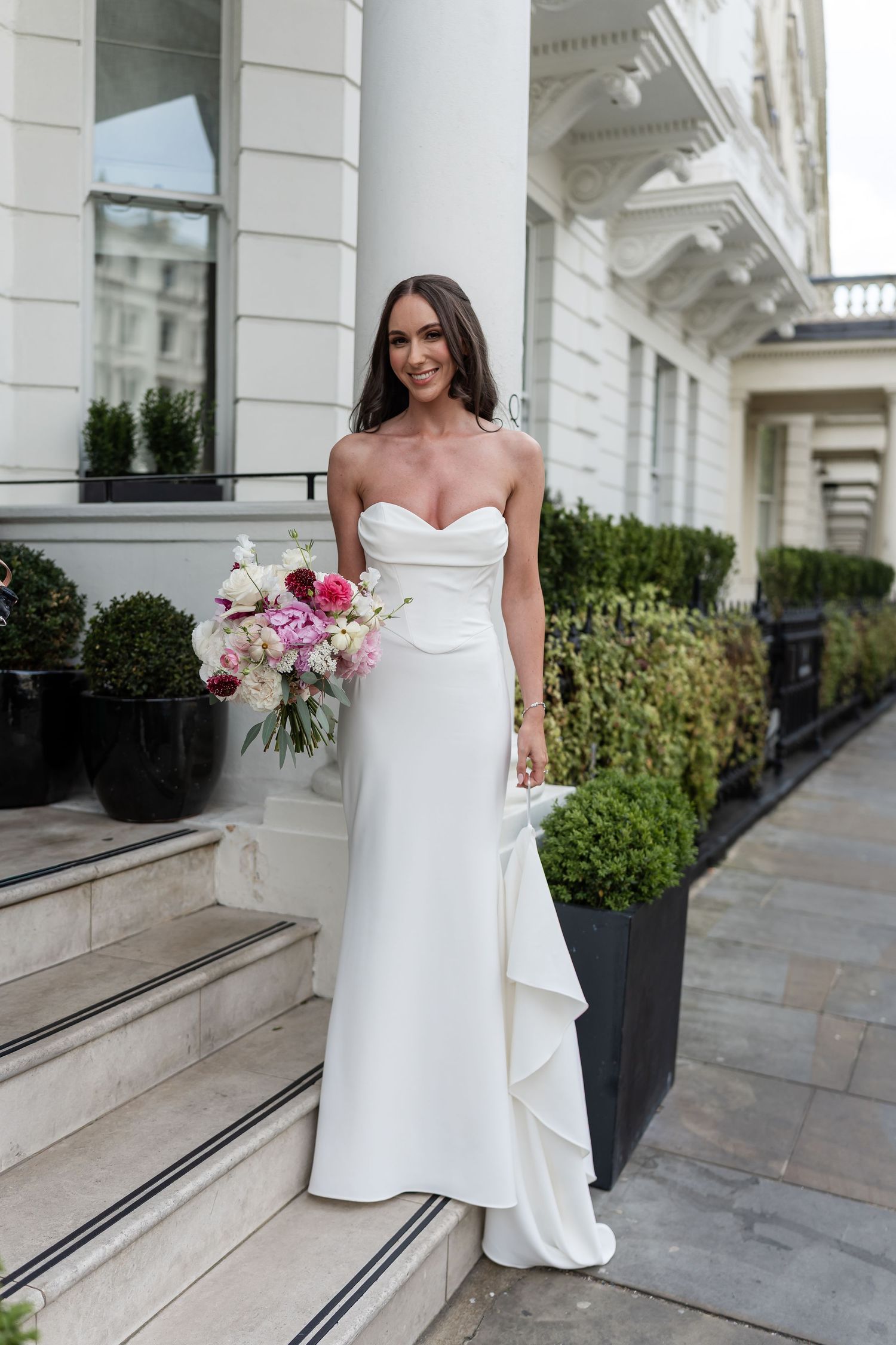 Elegant bride in strapless white gown holds pink bouquet on steps of white Georgian building.