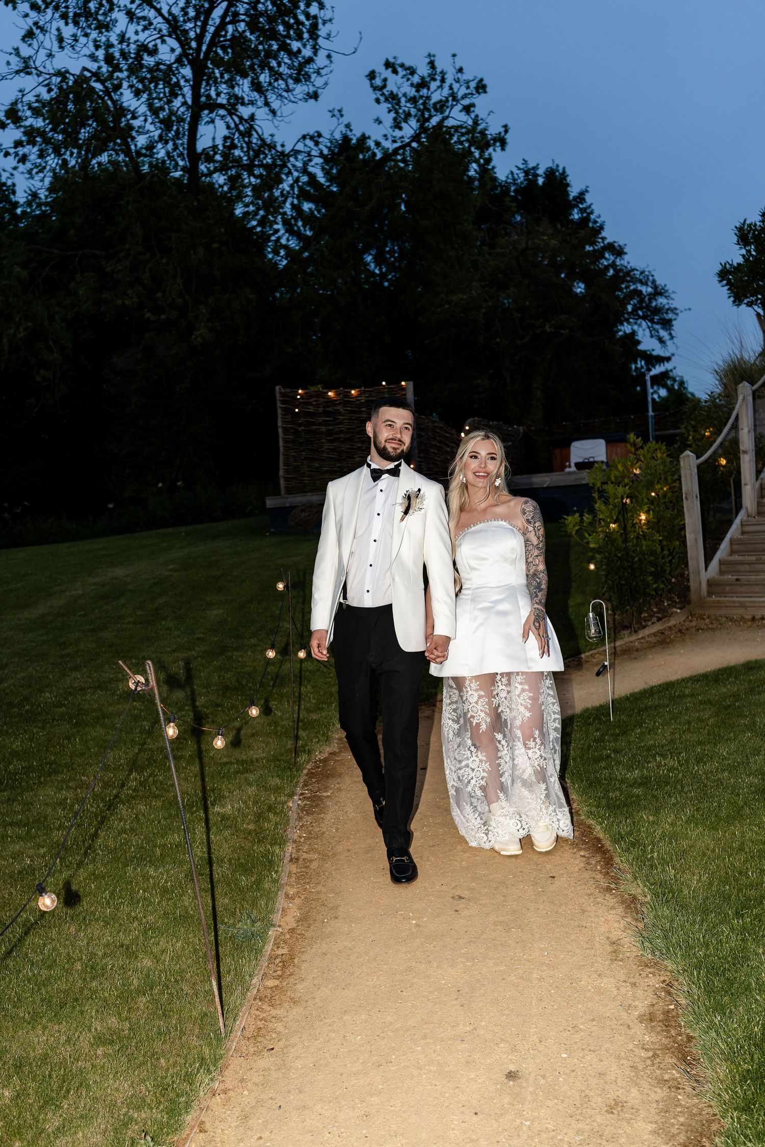 Newlywed couple walking hand in hand along a candlelit garden path at dusk in wedding attire.