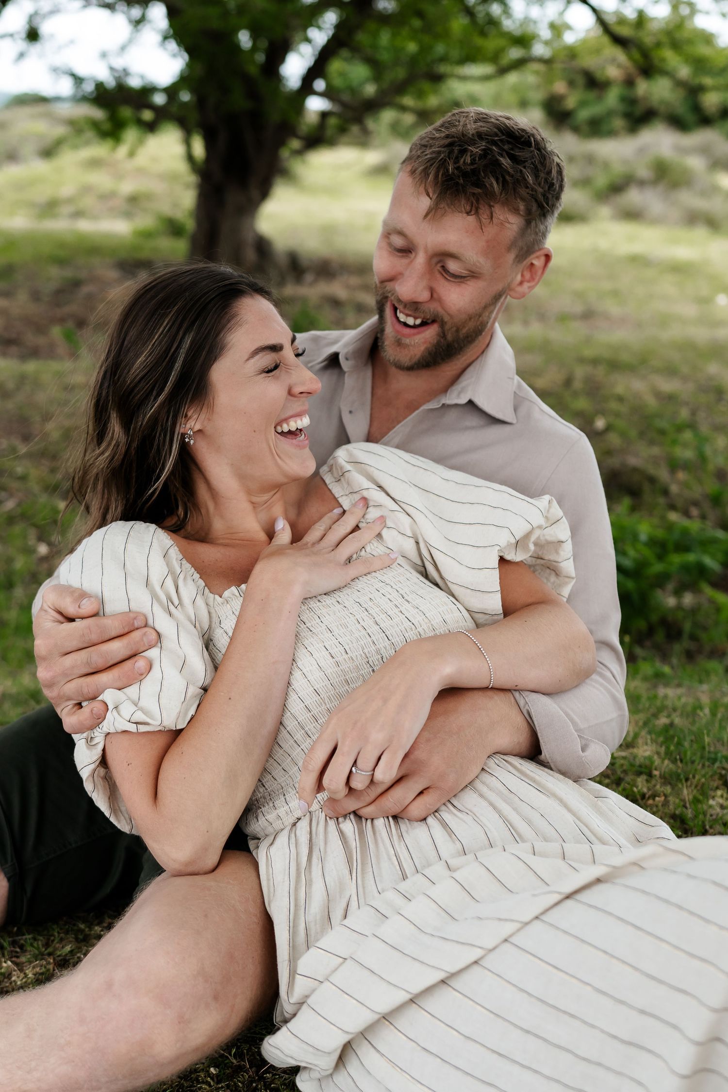 Joyful couple sitting on grass laughing together, bride-to-be and in the Norfolk countryside for their pre-wedding engagement photoshoot