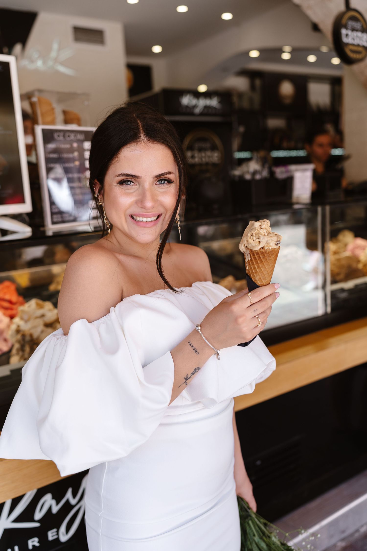 Radiant bride in white ruffled off-shoulder dress smiles holding a gelato cone inside an ice cream shop.