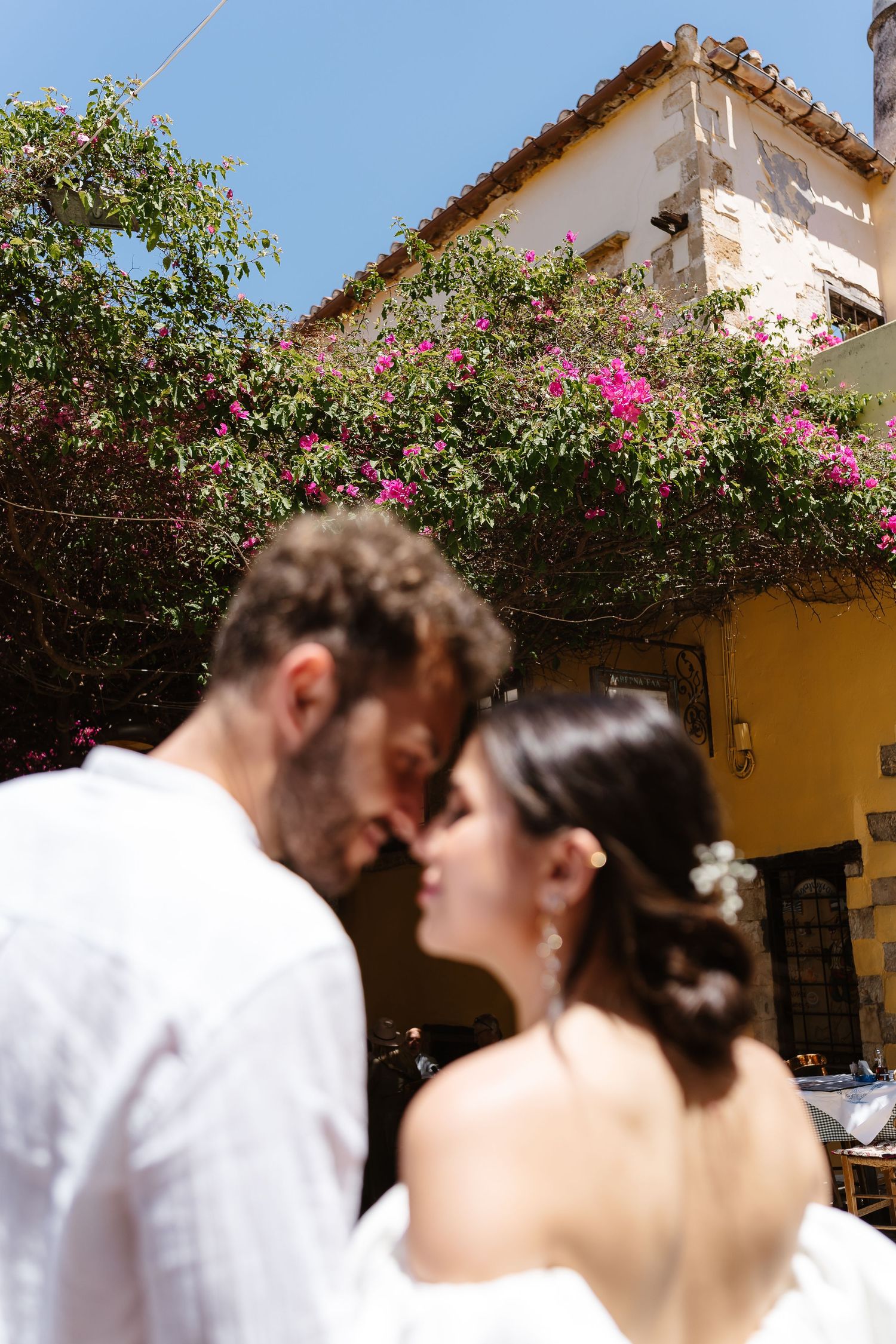 Couple in white attire touching foreheads romantically under blooming bougainvillea by a yellow Mediterranean building.