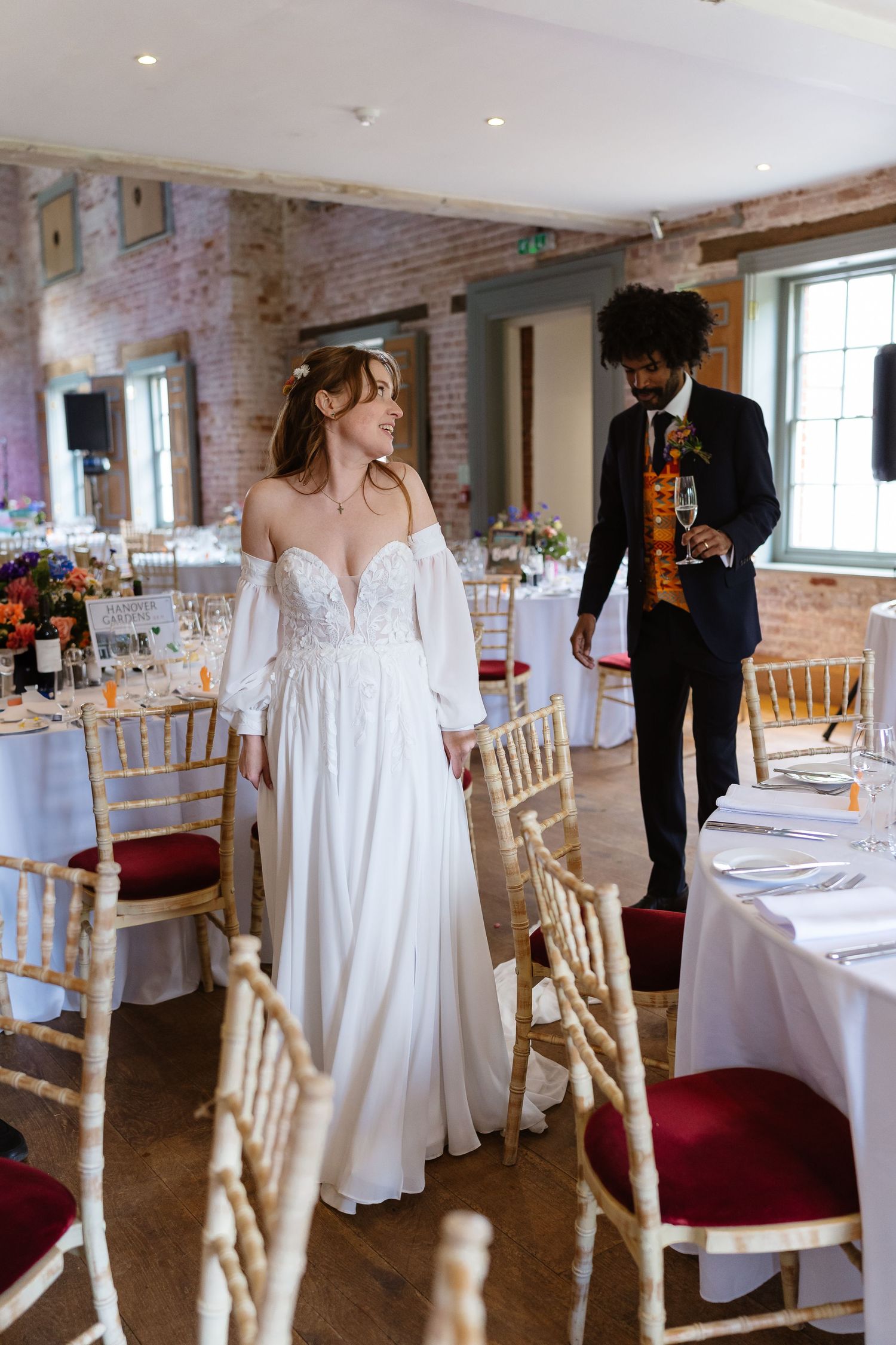 Bride in elegant white off-shoulder gown walks through decorated wedding reception hall with groom in navy suit behind her.