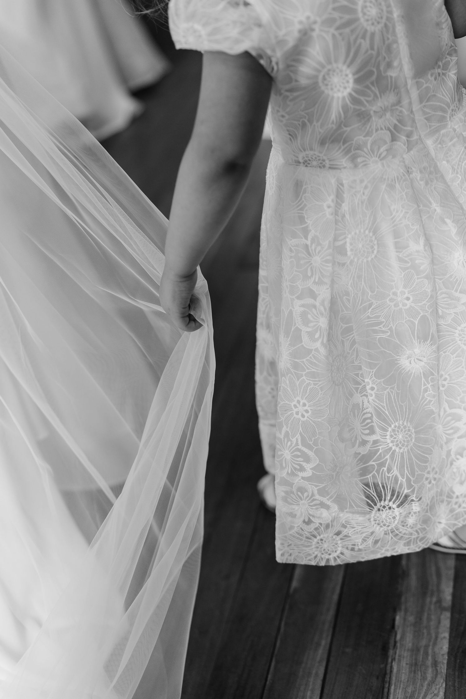 Black and white close-up of flower girl in lace dress holding brides tulle veil on wooden steps.