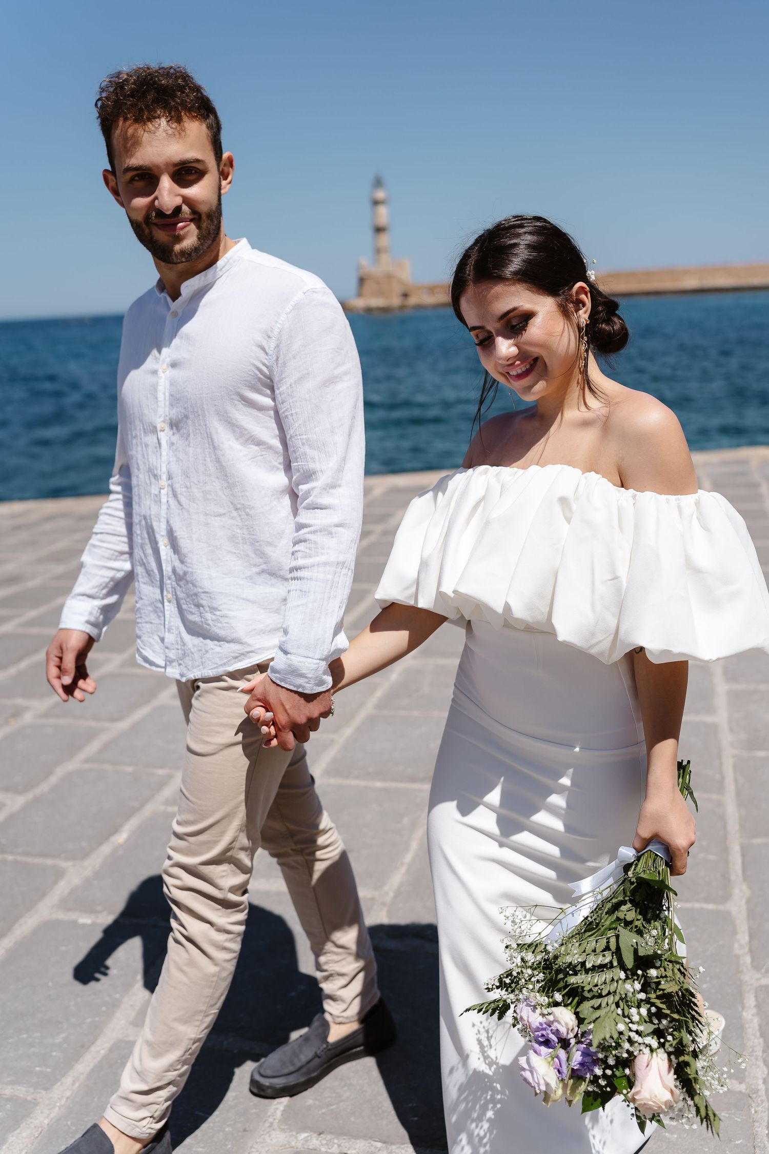 Smiling newlyweds walk hand-in-hand along a sunny Mediterranean waterfront, bride in white off-shoulder gown holding a bouquet.