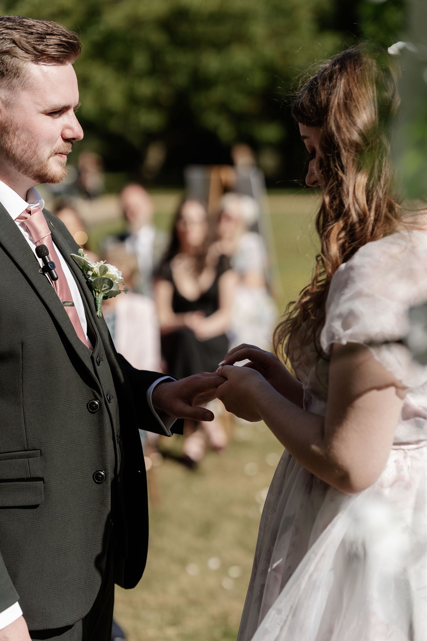 Bride places wedding ring on grooms finger during an outdoor ceremony with guests seated in background.