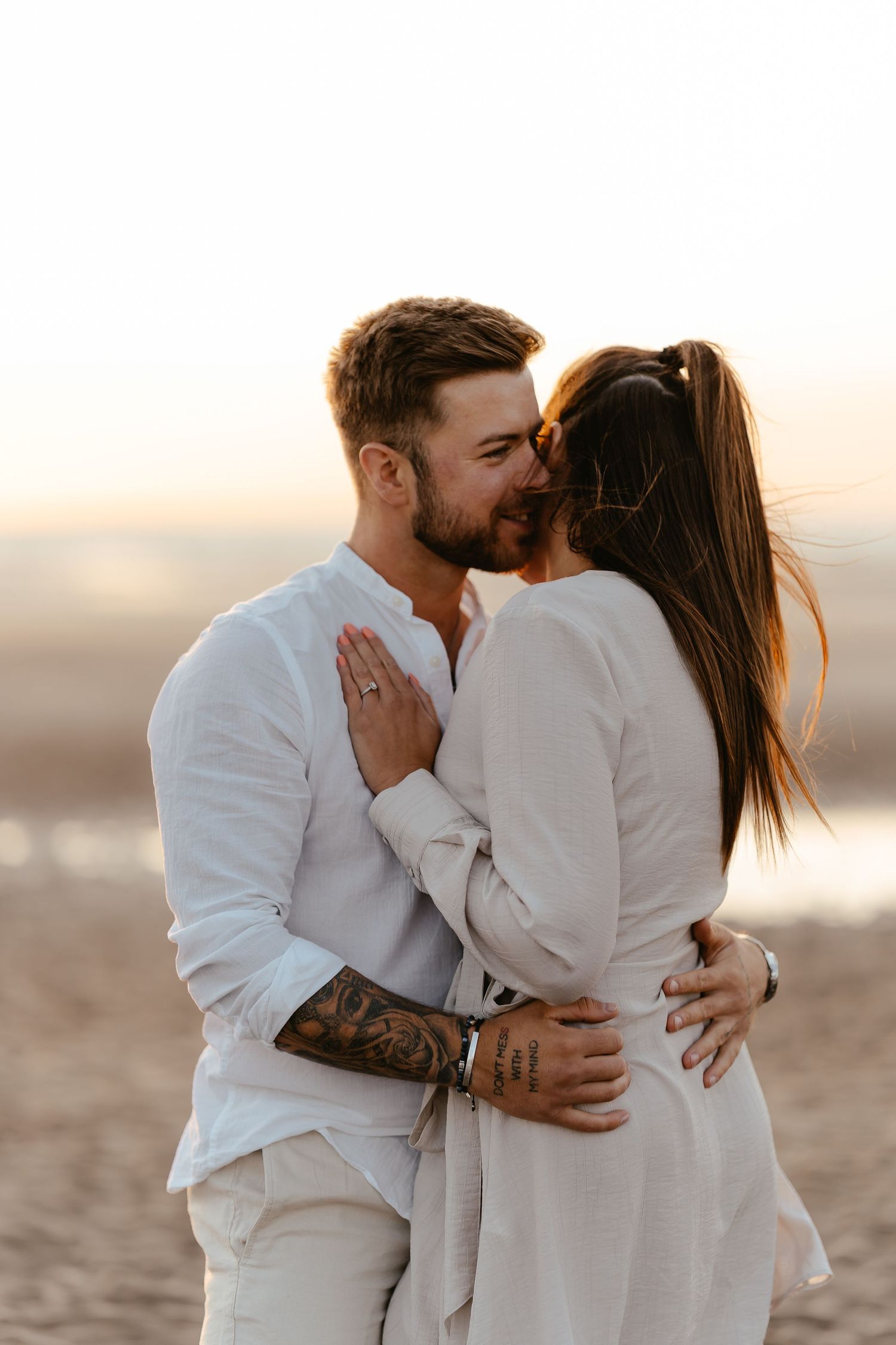 A couple sharing a romantic kiss on a sandy beach at golden sunset, both dressed in white linen outfits.