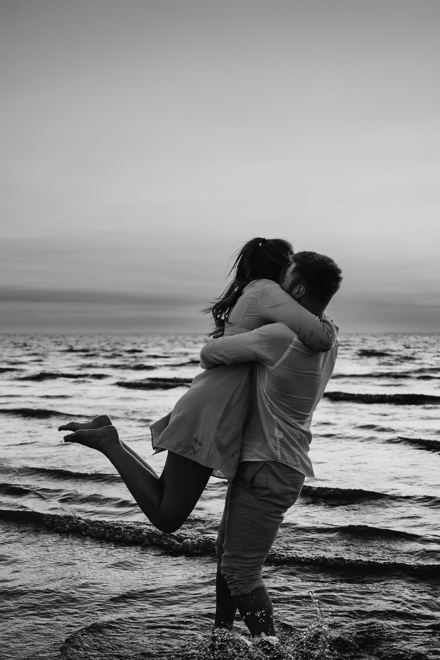 Black and white photo of couple embracing on beach shoreline at dusk.