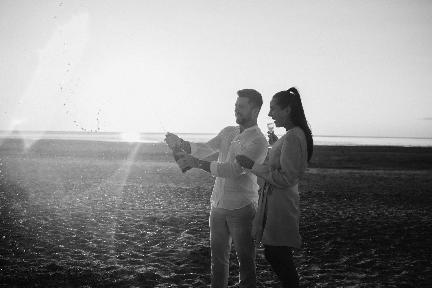 Black and white image of a couple spraying champagne on a sunlit beach, laughing together.