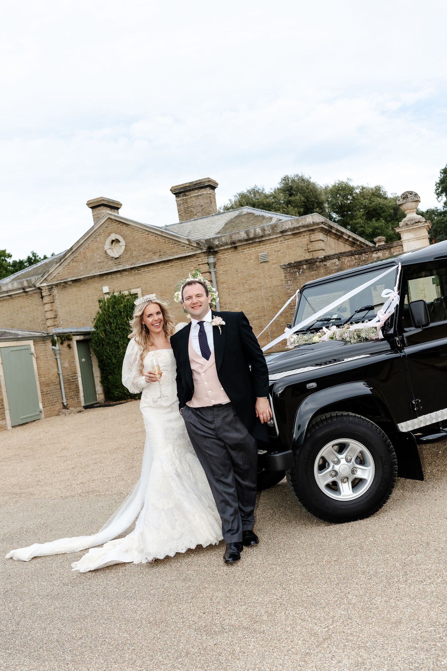 Bride and groom posing beside decorated black Land Rover Defender outside rustic wedding venue.