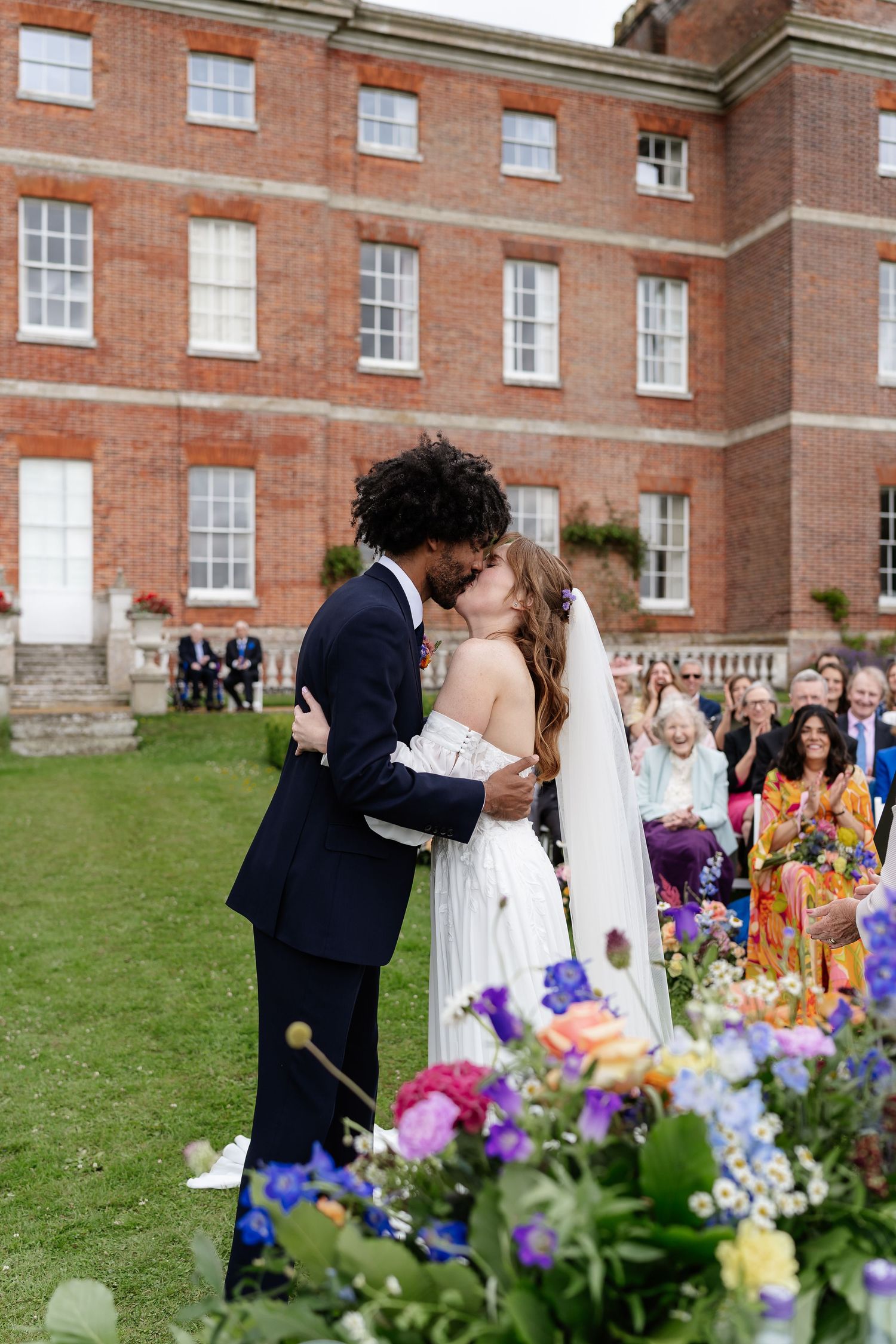 Newlywed couple shares first kiss in front of grand red brick manor surrounded by colorful floral arrangements.