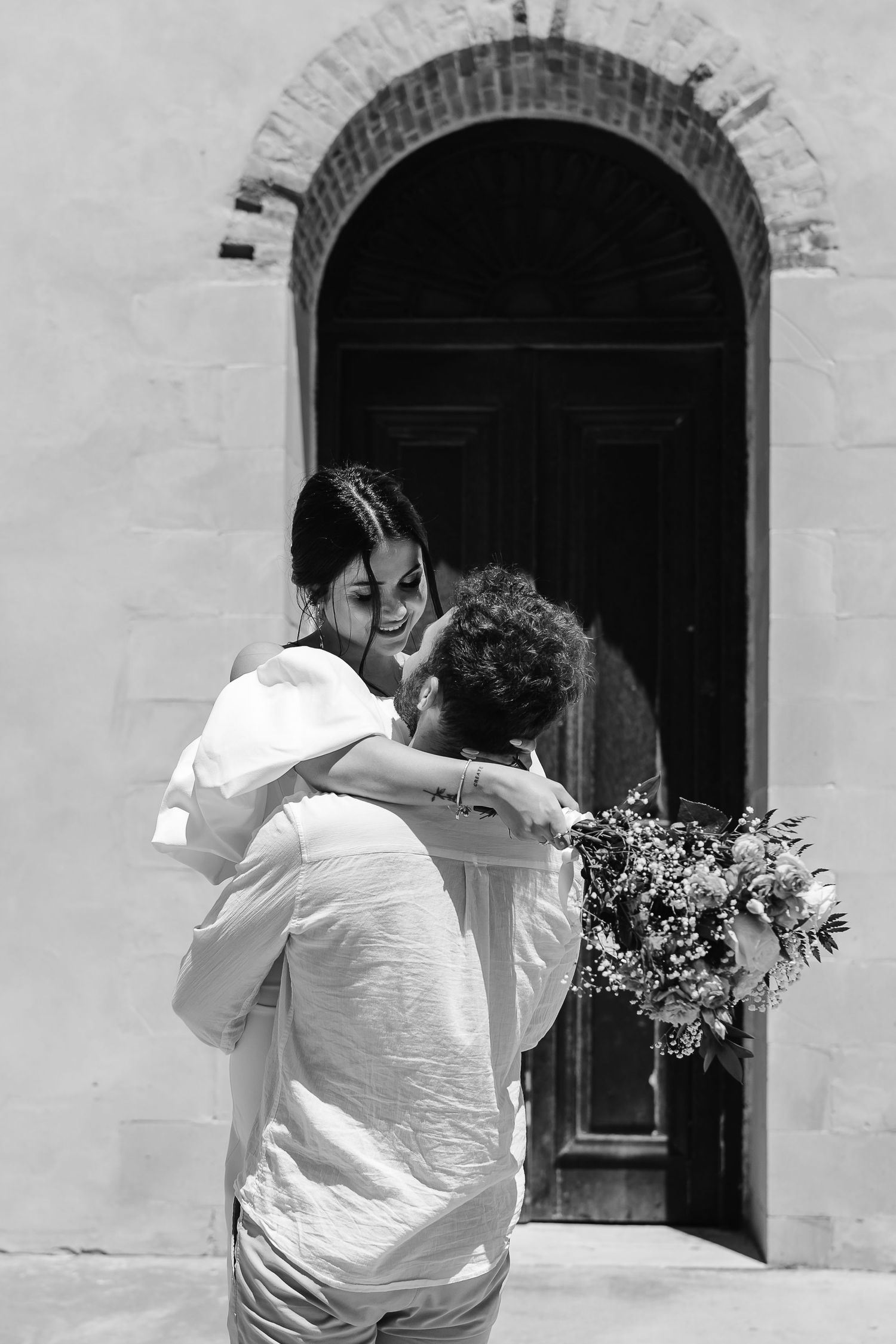 Black and white photo of groom lifting bride near an arched wooden door, bride holding a wildflower bouquet overhead.