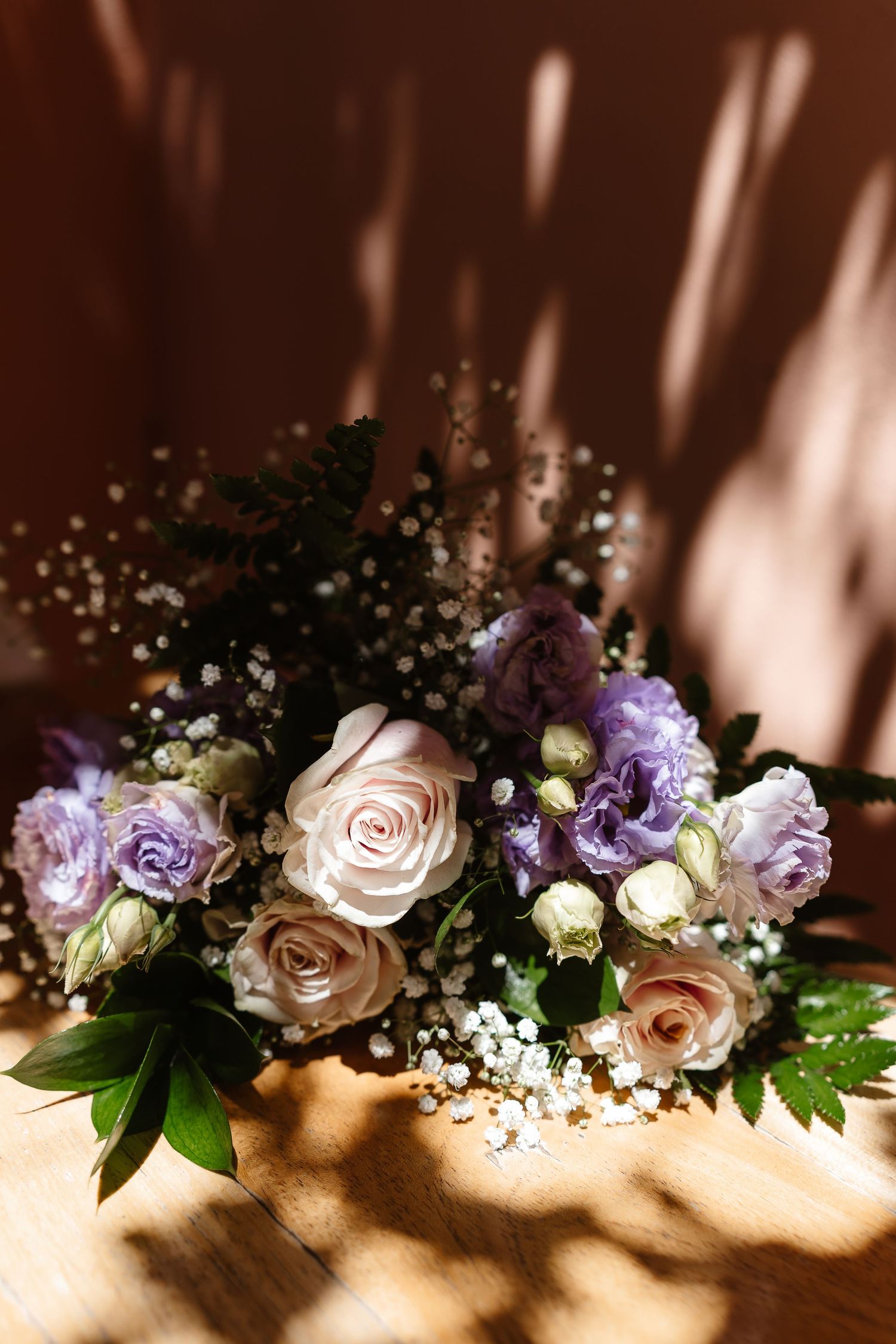 Elegant wedding bouquet of purple and cream roses with gypsophila on a wooden table in warm sunlight.