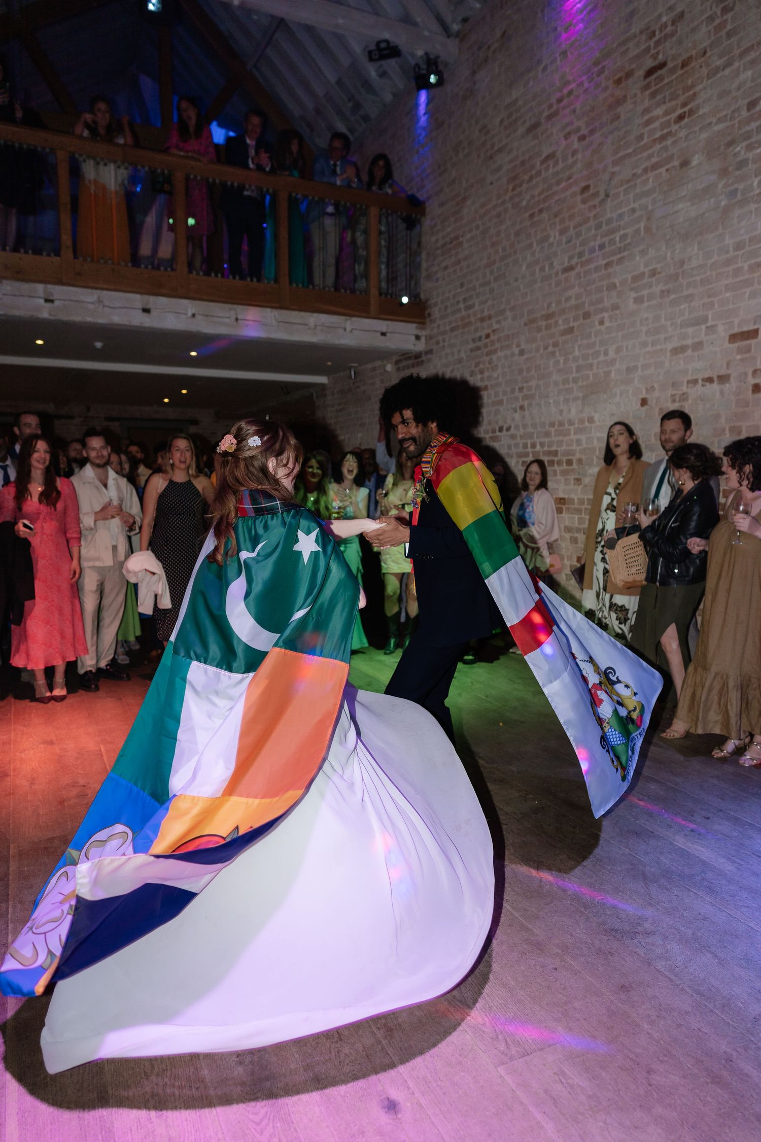 Newlyweds twirl displaying large international flag capes on wedding reception dance floor.