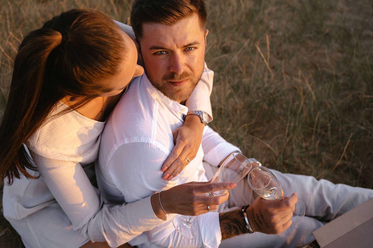 Couple clinking champagne glasses during a golden hour picnic, woman affectionately leaning on mans shoulder.