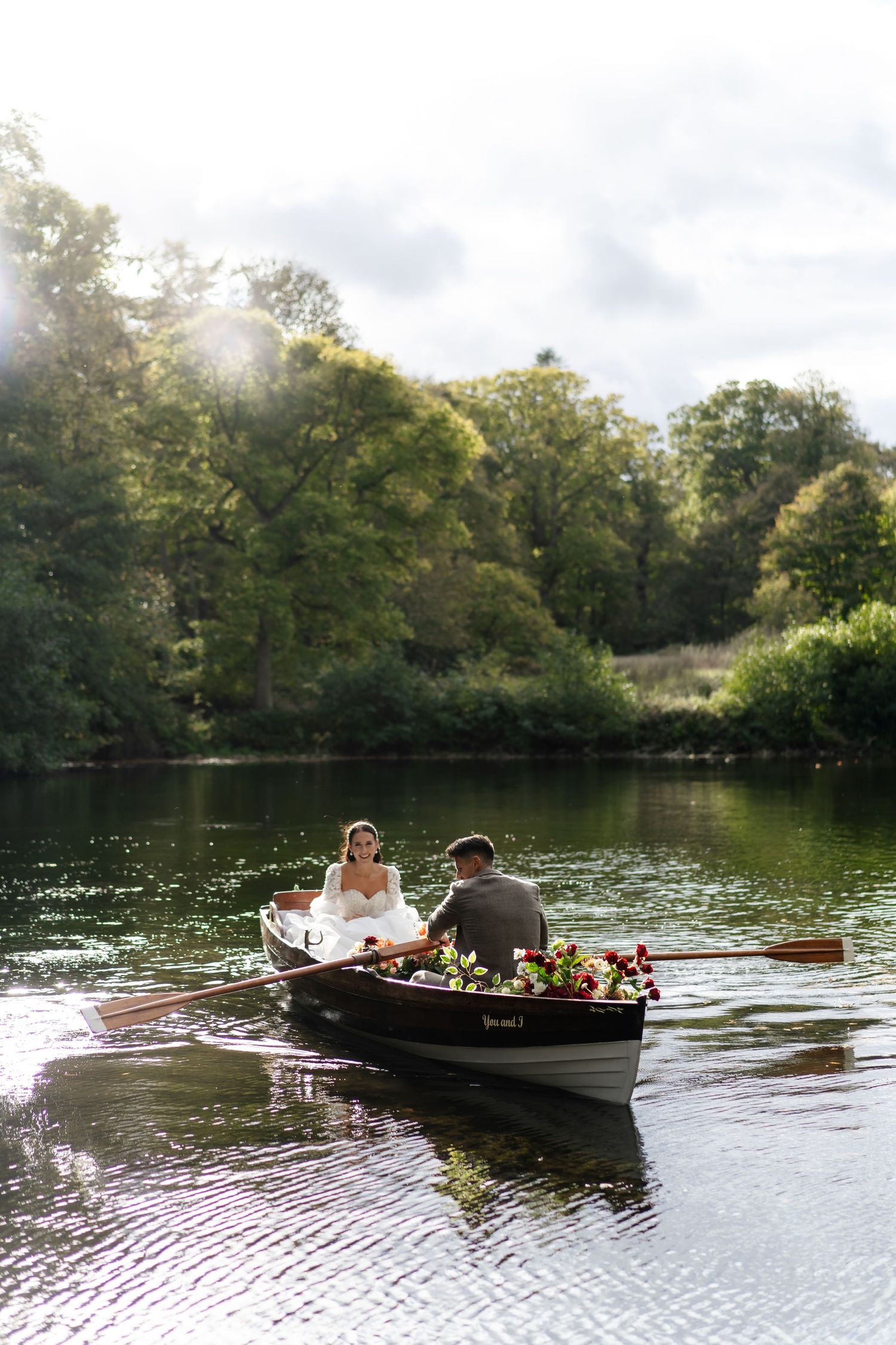 Bride and groom sharing a romantic rowboat ride on a sunlit river surrounded by lush trees.