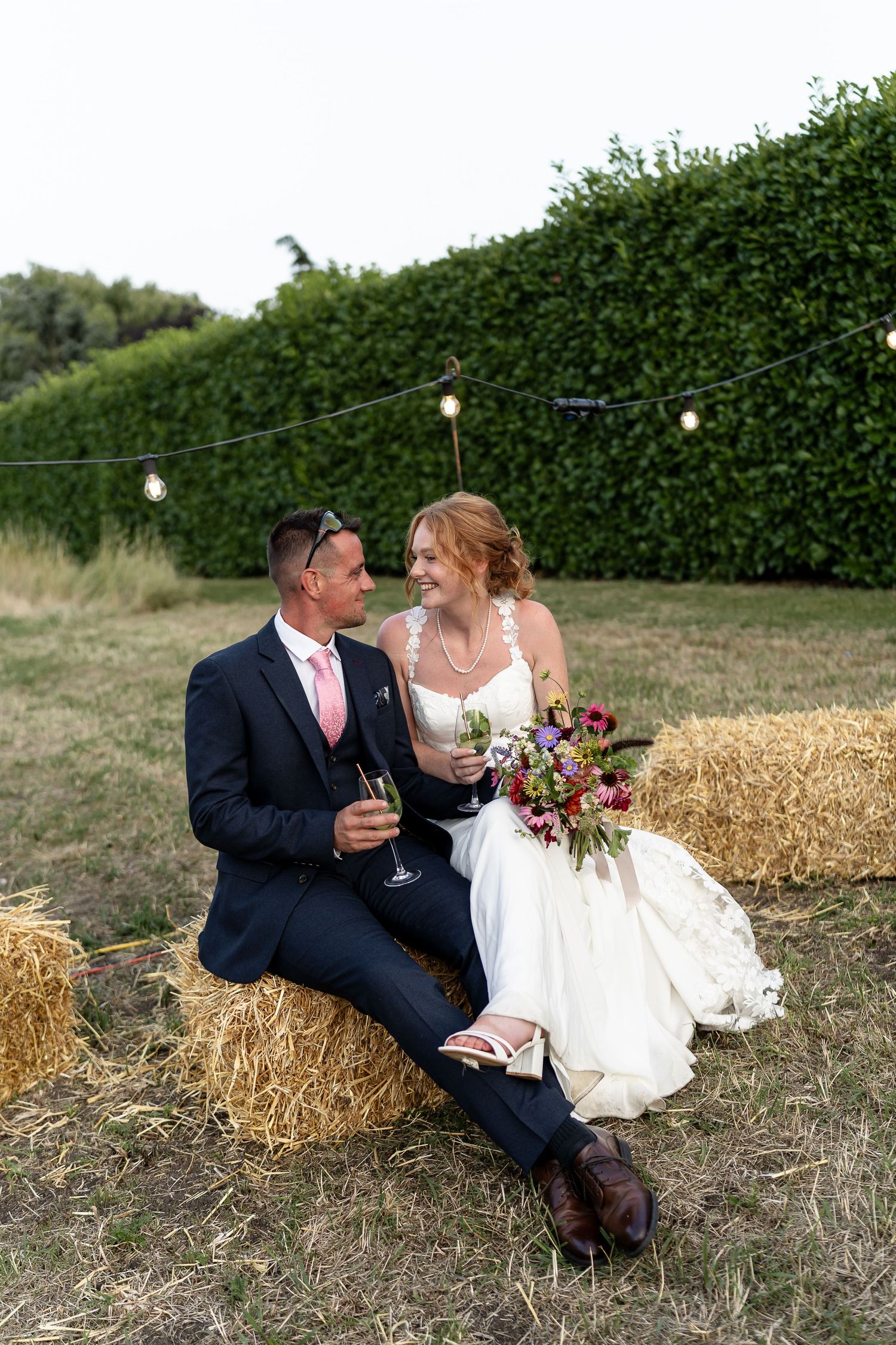 Bride and groom sitting on hay bales holding champagne glasses with colorful bouquet at outdoor wedding reception.