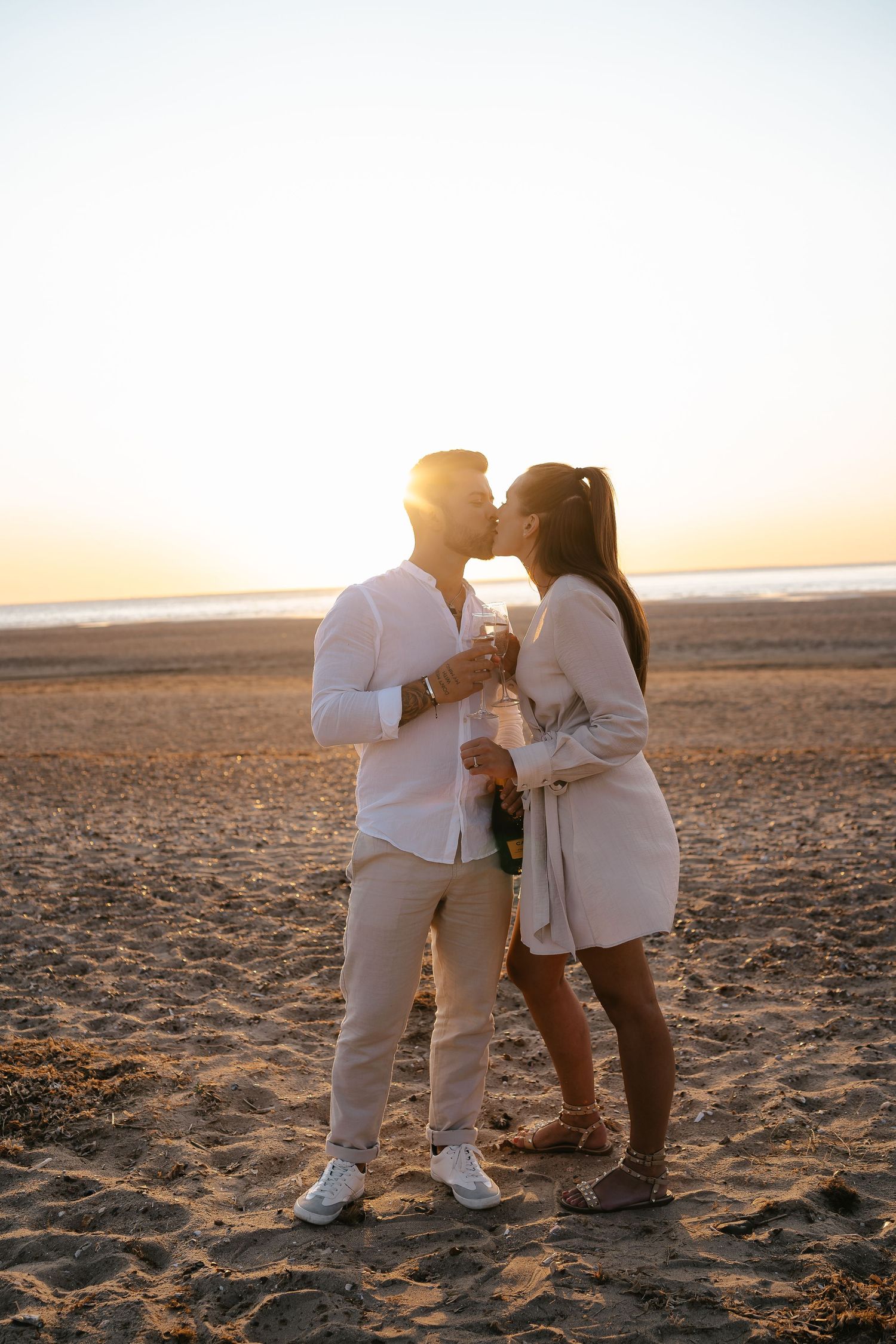 Romantic couple kissing on a beach at sunset while holding champagne glasses and a bottle.
