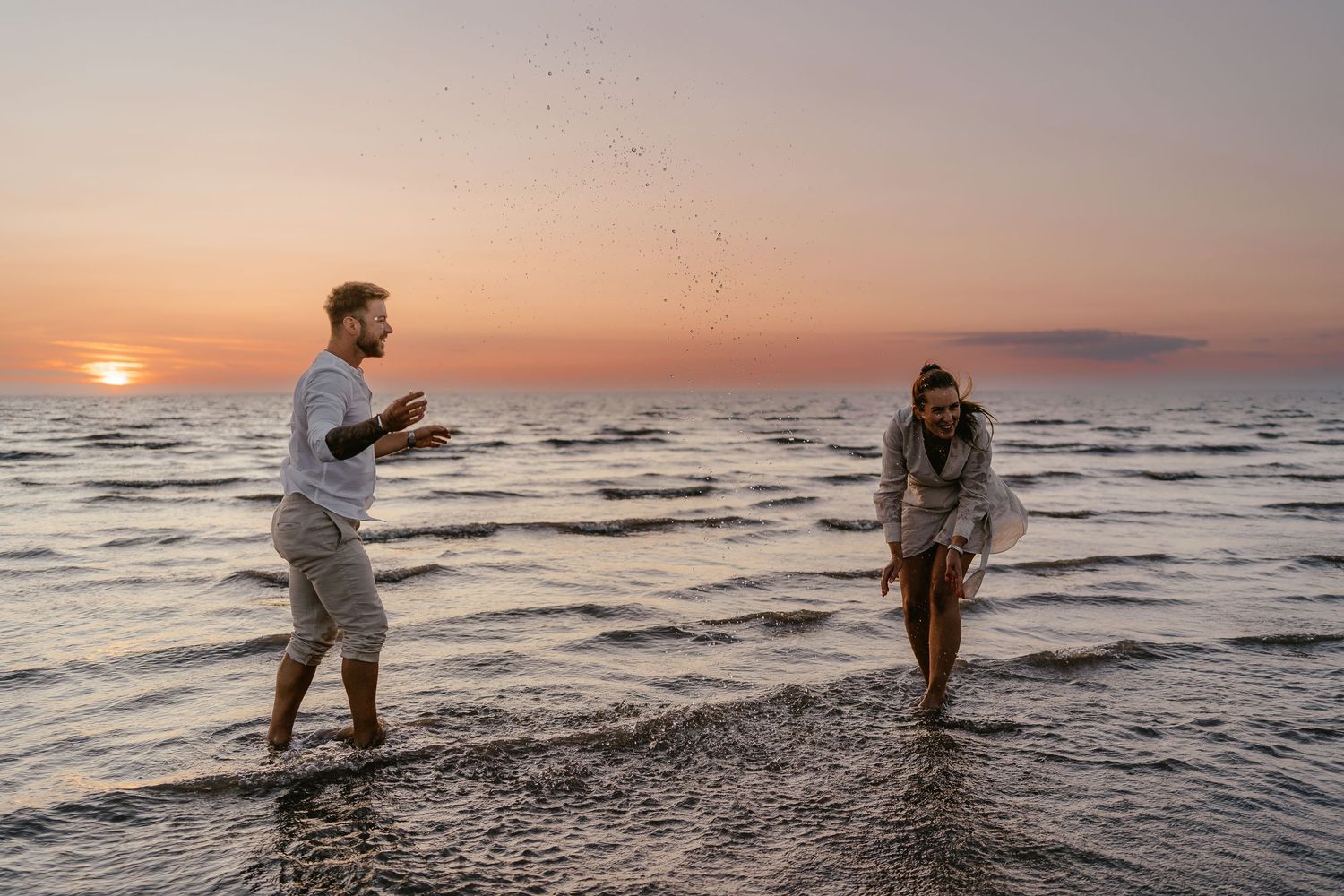 Couple playfully splashing water at each other on beach during golden sunset.