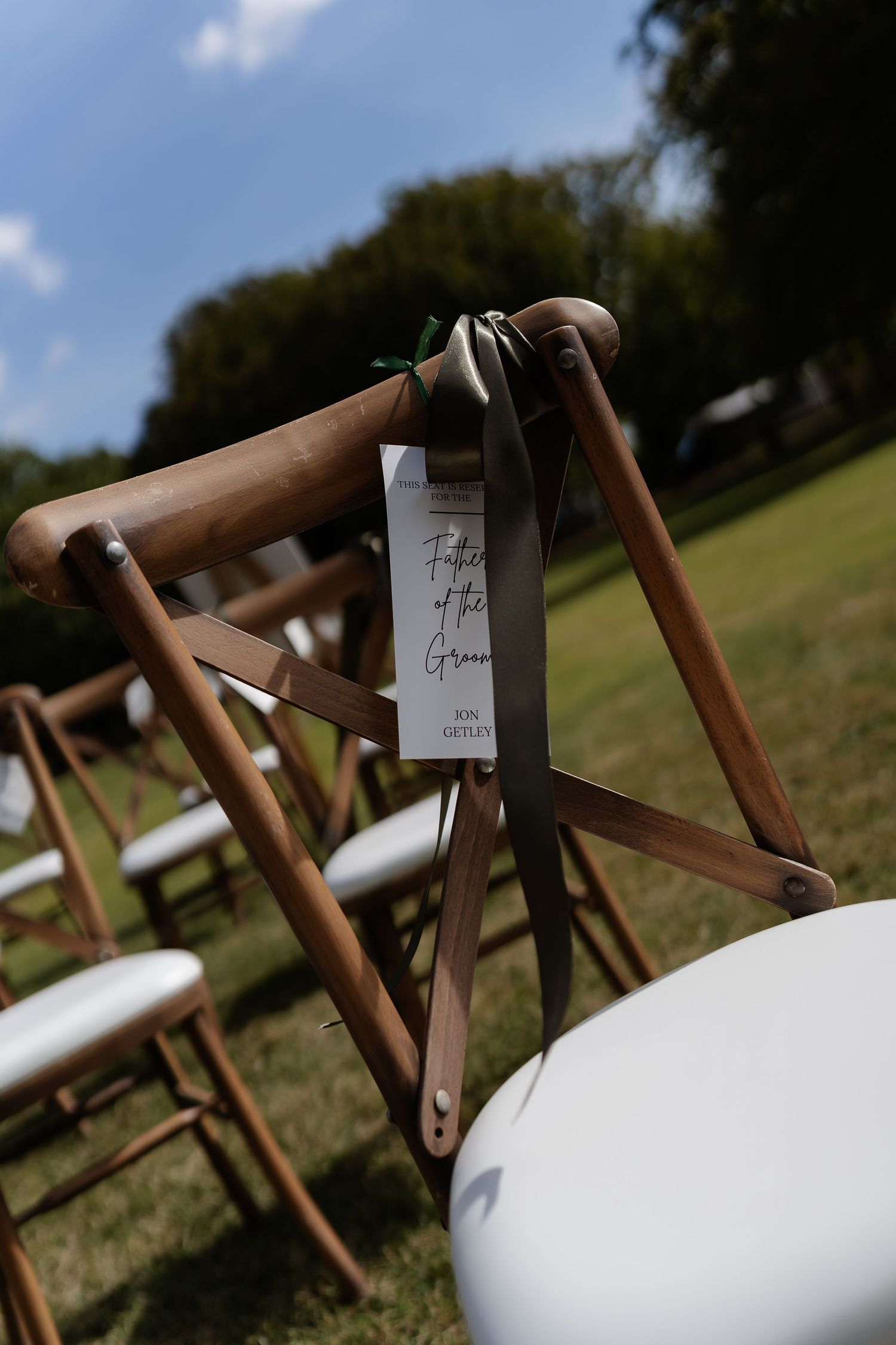 Wooden folding chair with a Father of the Groom reserved sign tied with sage green ribbon at an outdoor ceremony.