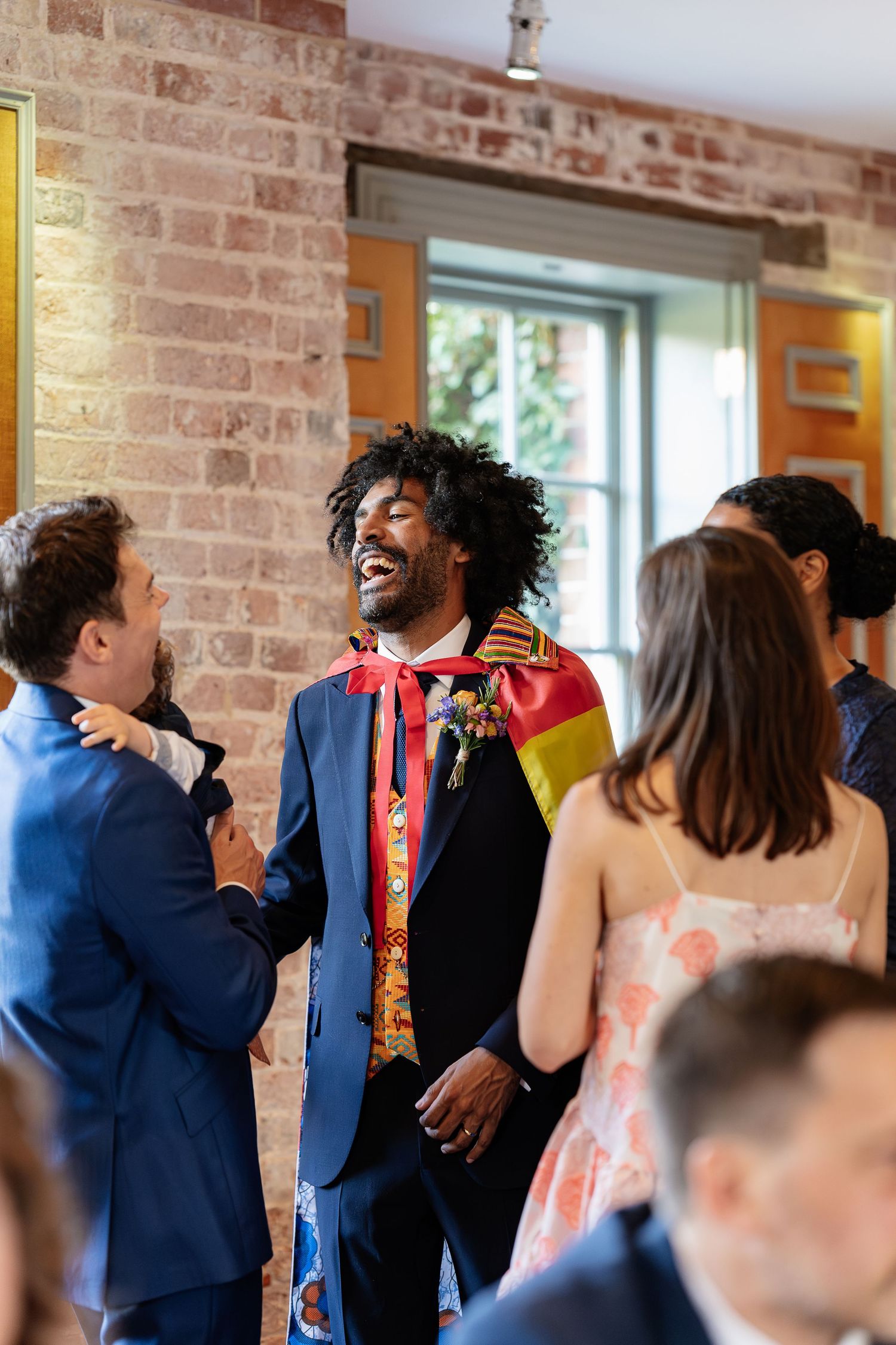 Groom in colorful ceremonial jacket laughs joyfully while socializing with guests at a rustic brick-walled wedding reception.