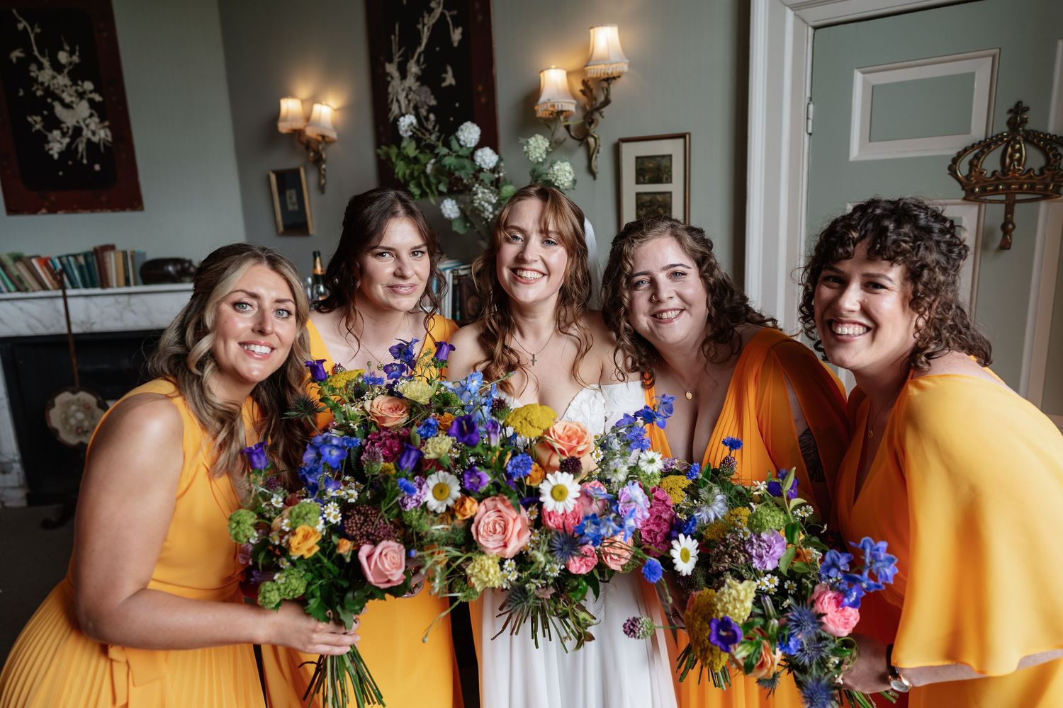 Bride with four bridesmaids in yellow dresses holding colorful wildflower bouquets smiling together indoors.