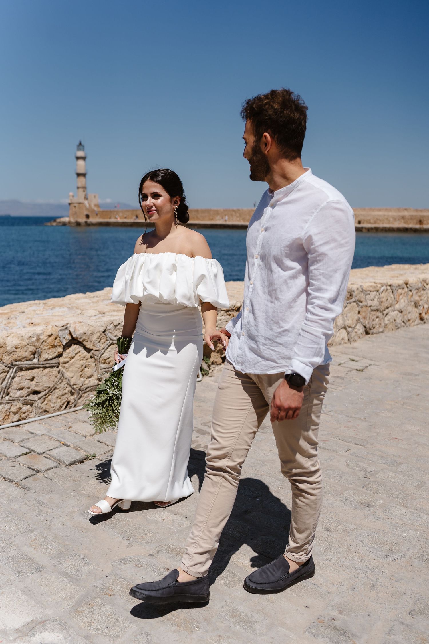 Smiling bride and groom holding hands strolling along Chania harbor with lighthouse backdrop in Crete.