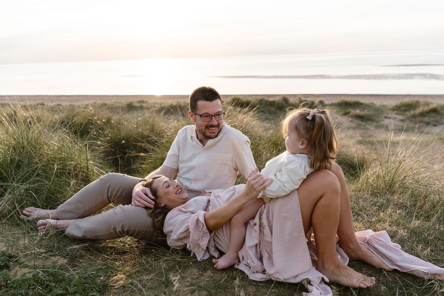 Family of three relaxes together in coastal grass dunes at sunset by the seaside.