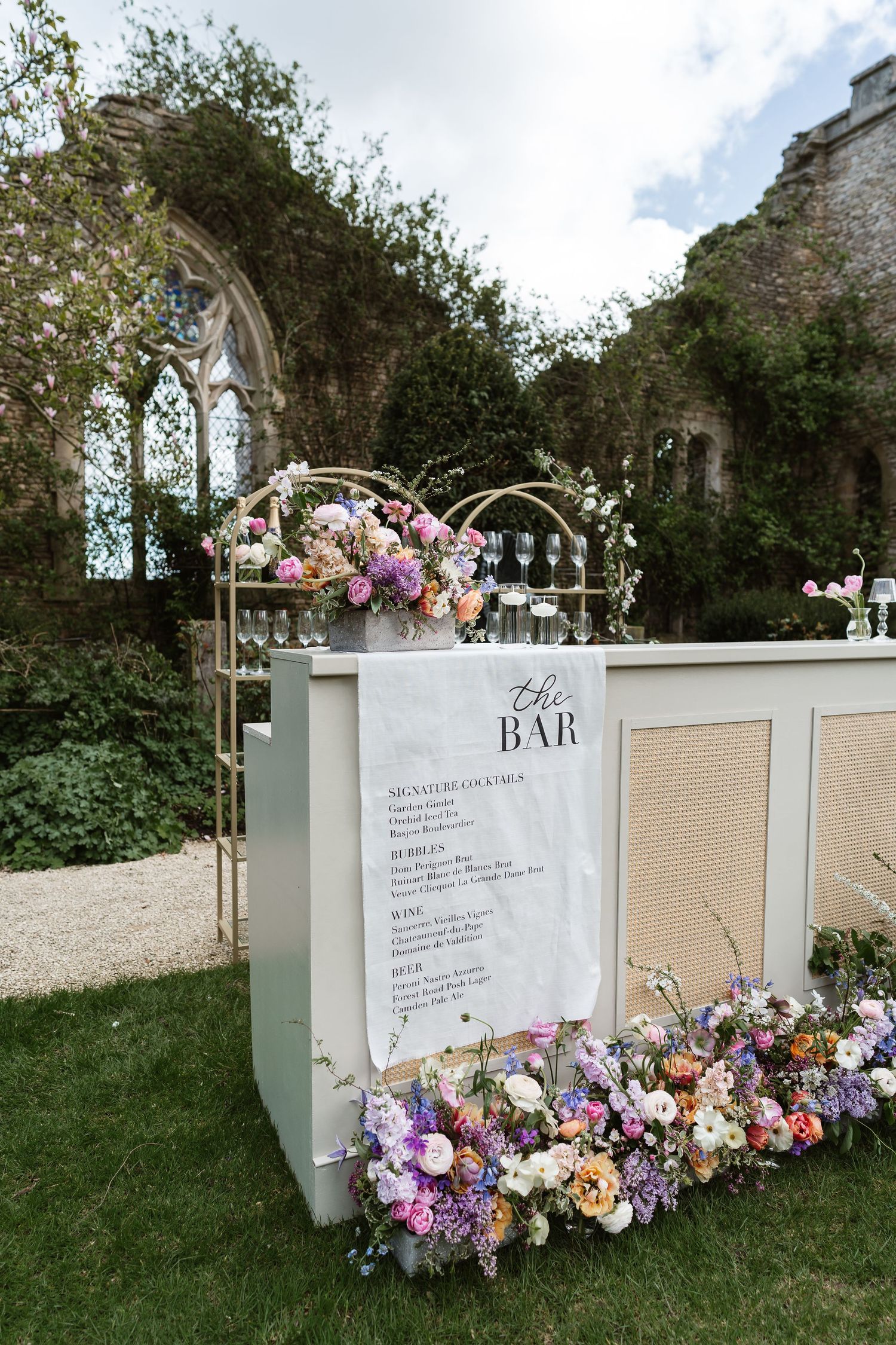 Elegant outdoor wedding bar decorated with lush colorful florals set against a rustic stone arch backdrop.
