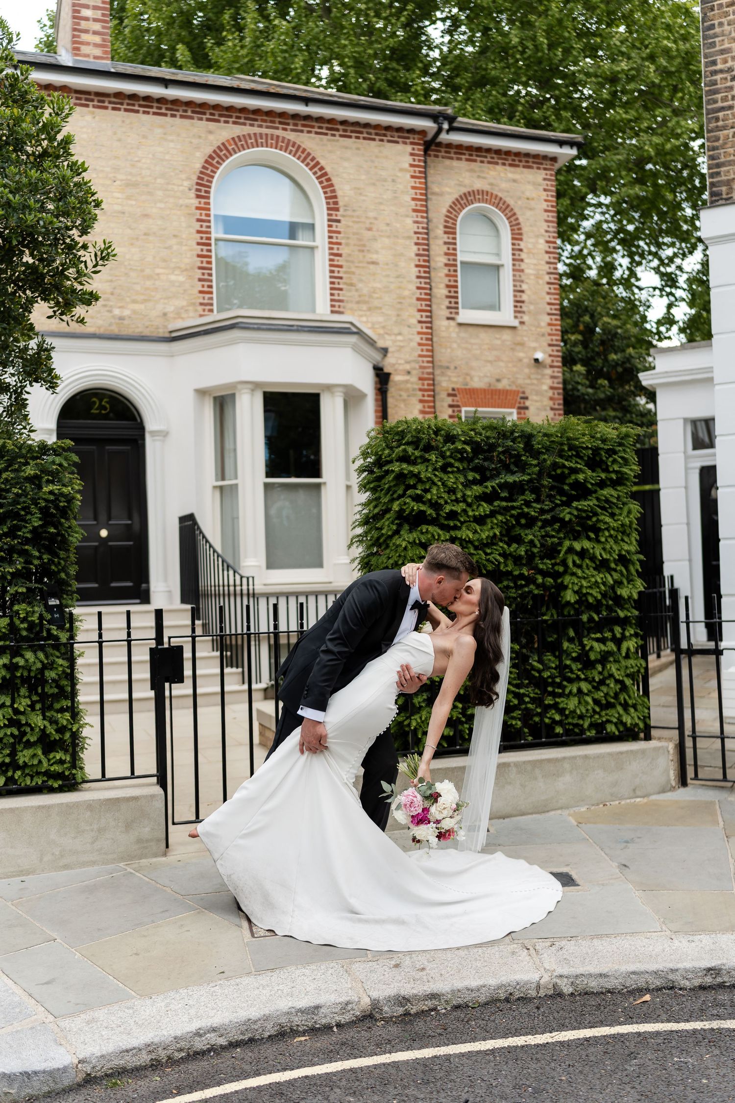 Bride and groom sharing romantic dip kiss on London street corner in elegant wedding attire with bridal bouquet.