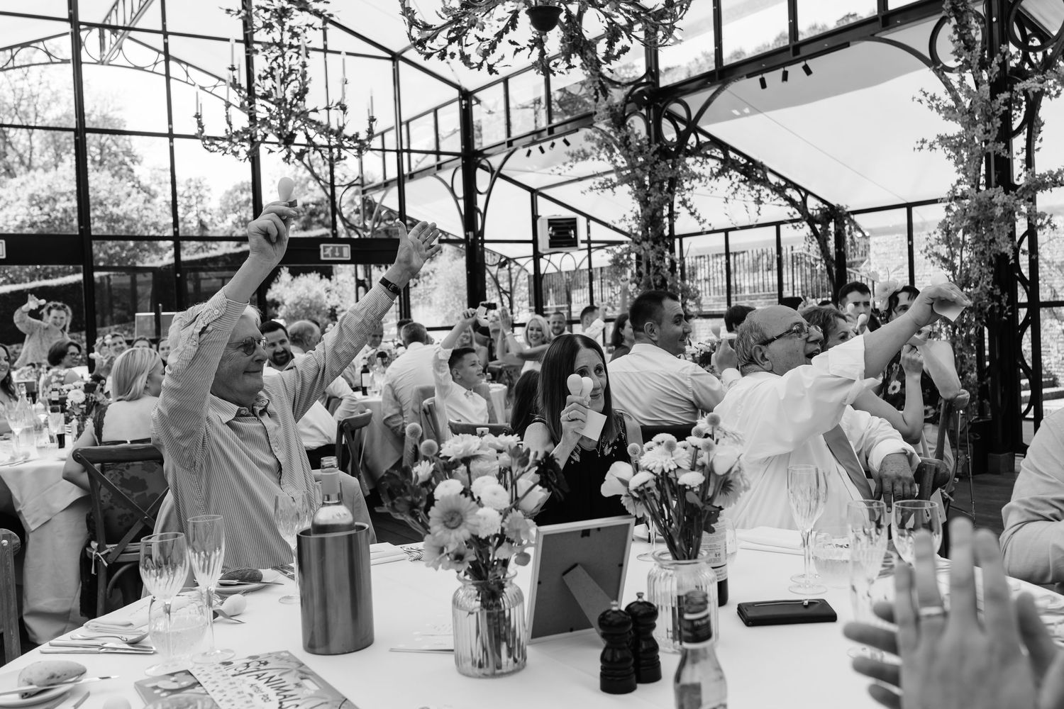 Black and white photo of wedding reception guests raising glasses in a toast inside a greenhouse venue.