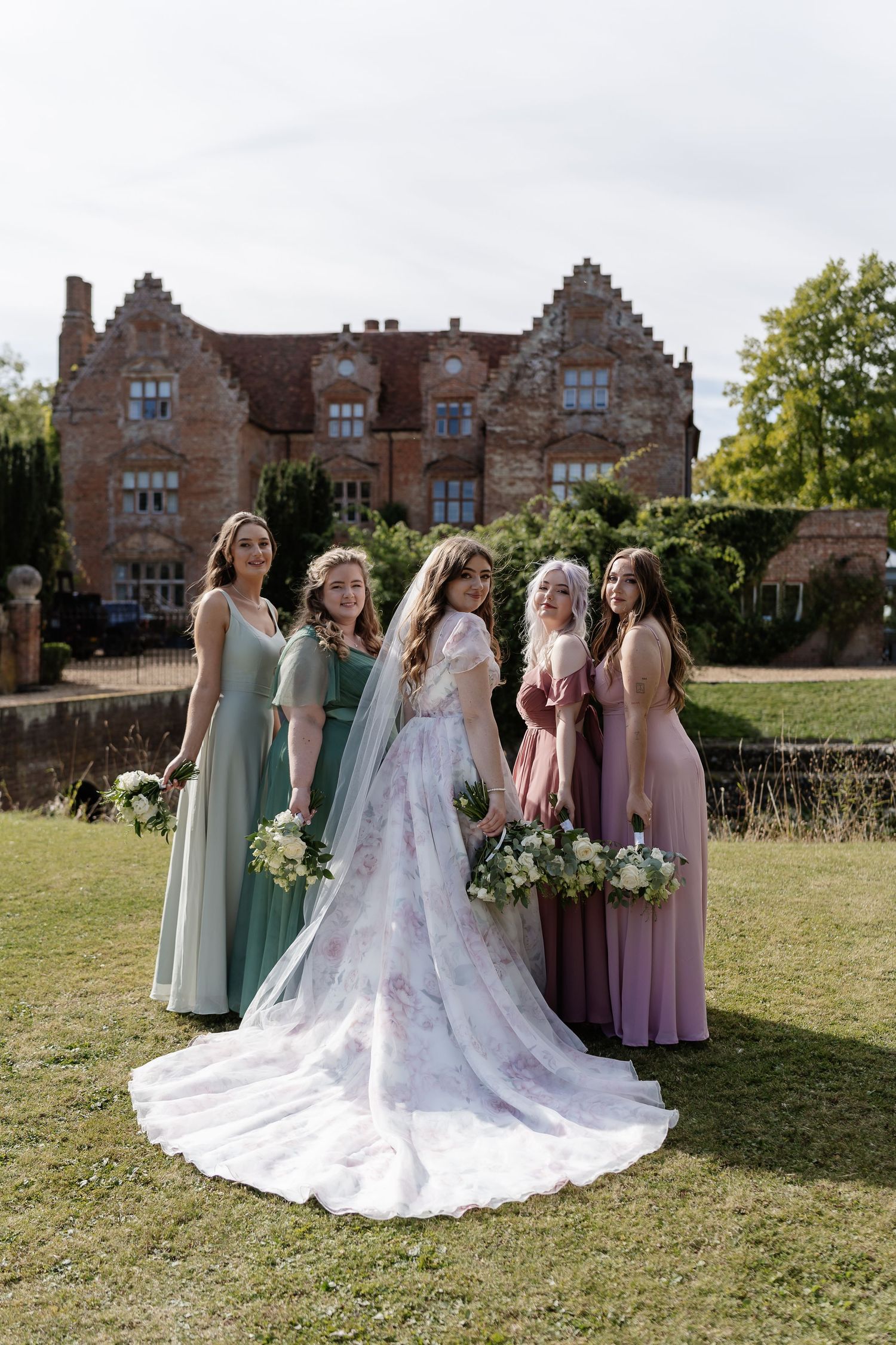 Bride in floral wedding gown stands with bridesmaids in green and pink dresses outside an elegant manor.