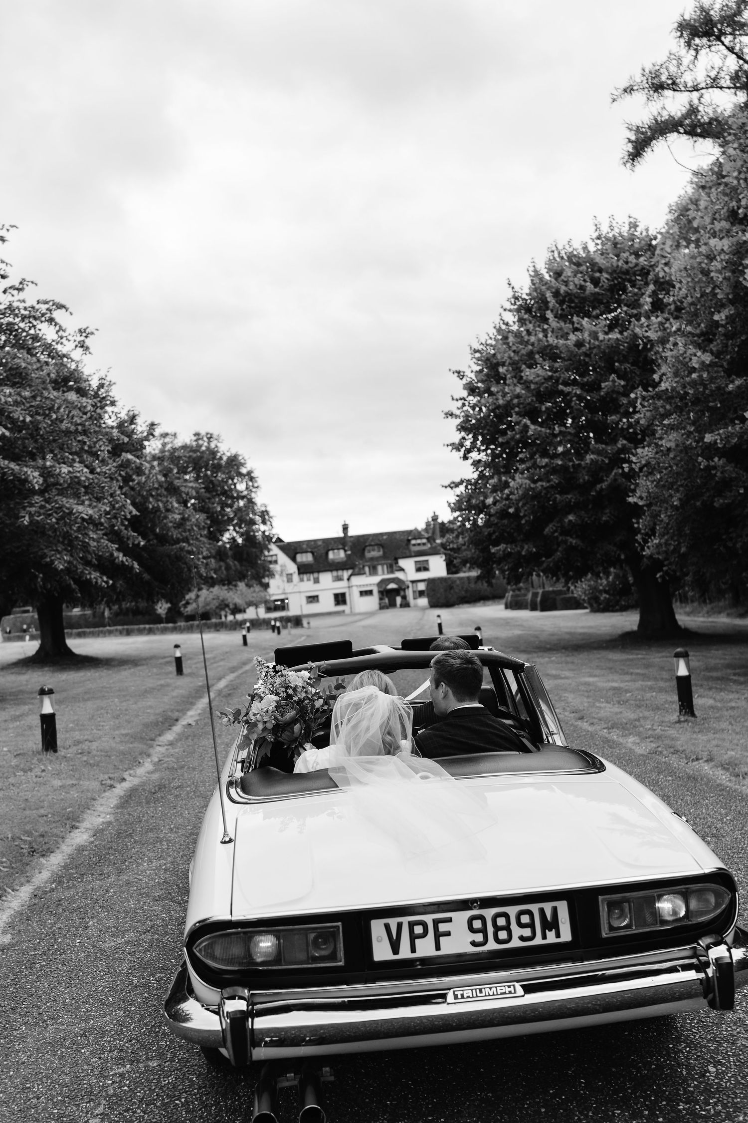 Black and white photo of a bride arriving in a vintage convertible car at her wedding venue.