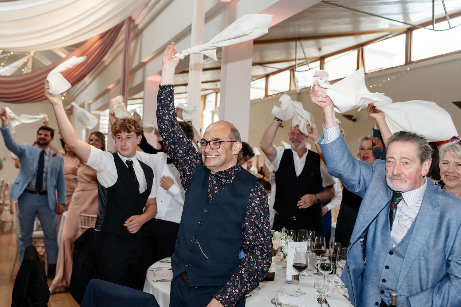 Joyful wedding reception guests wave white napkins in the air during celebratory dinner toast.