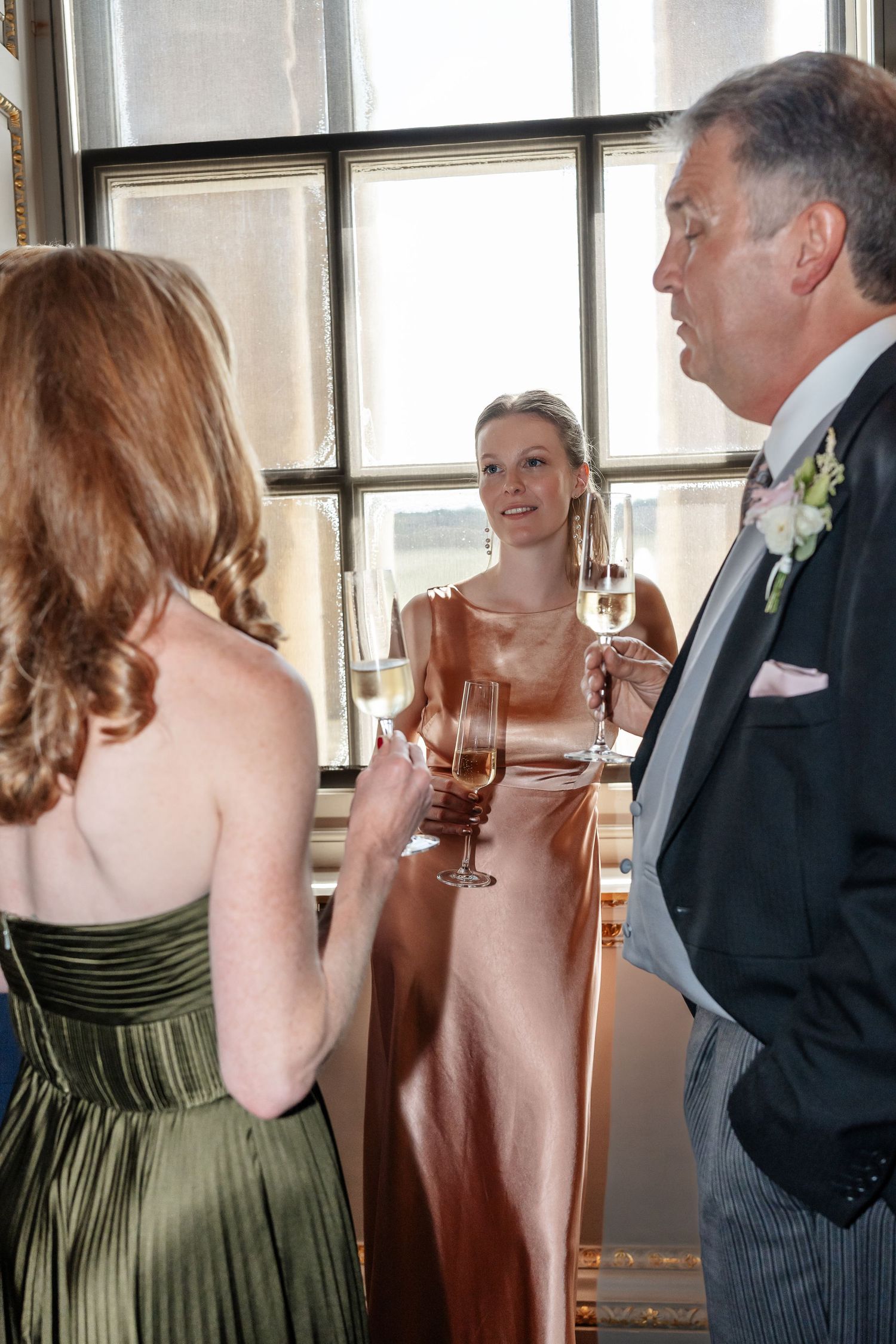 Wedding guests in formal attire chat and enjoy champagne near large windows at elegant reception venue.
