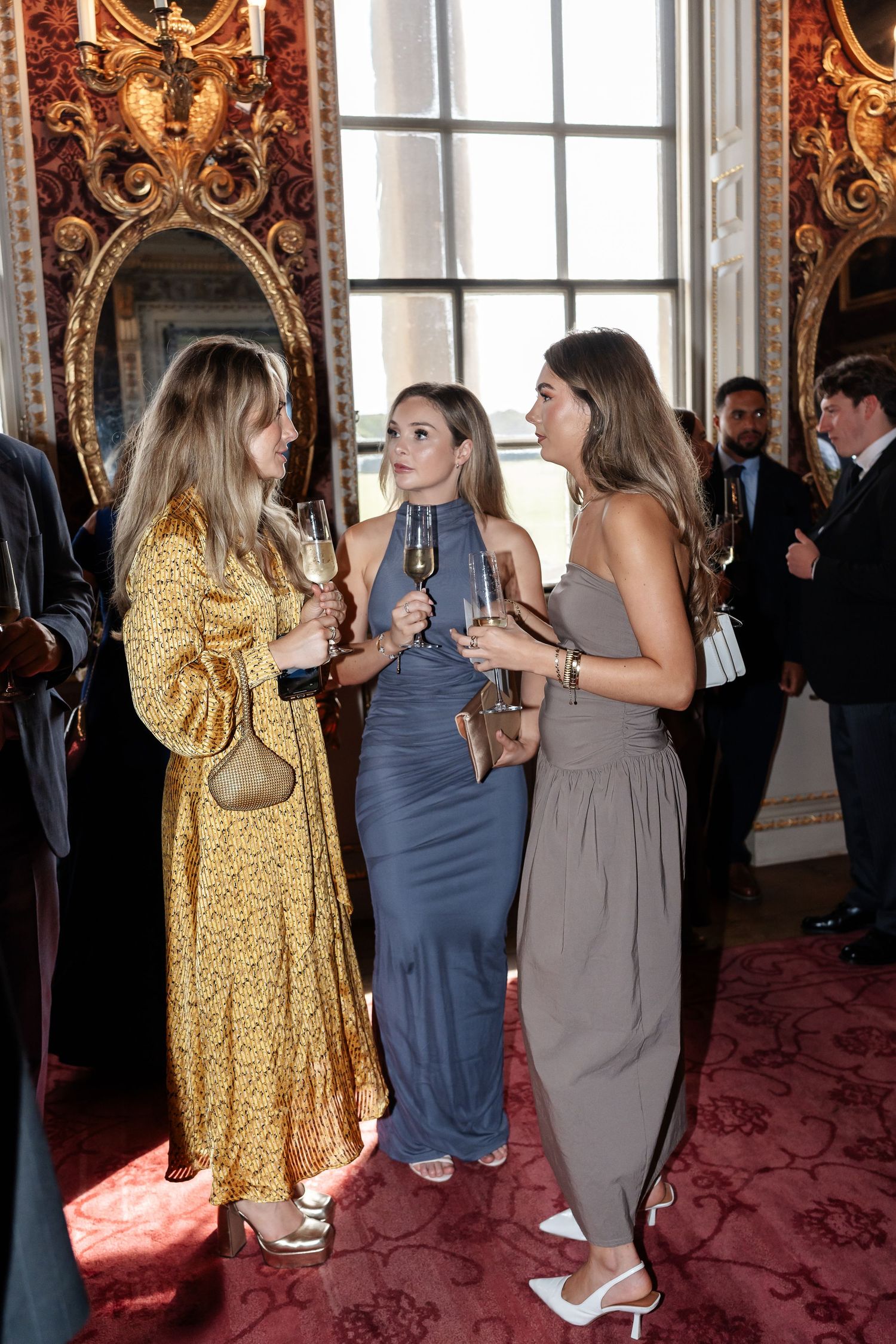 Three elegantly dressed wedding guests socializing and holding champagne glasses in ornate reception room.