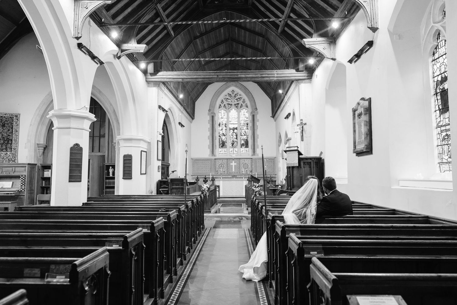 Black and white photo of bride and groom sitting in historic church pew before the altar.