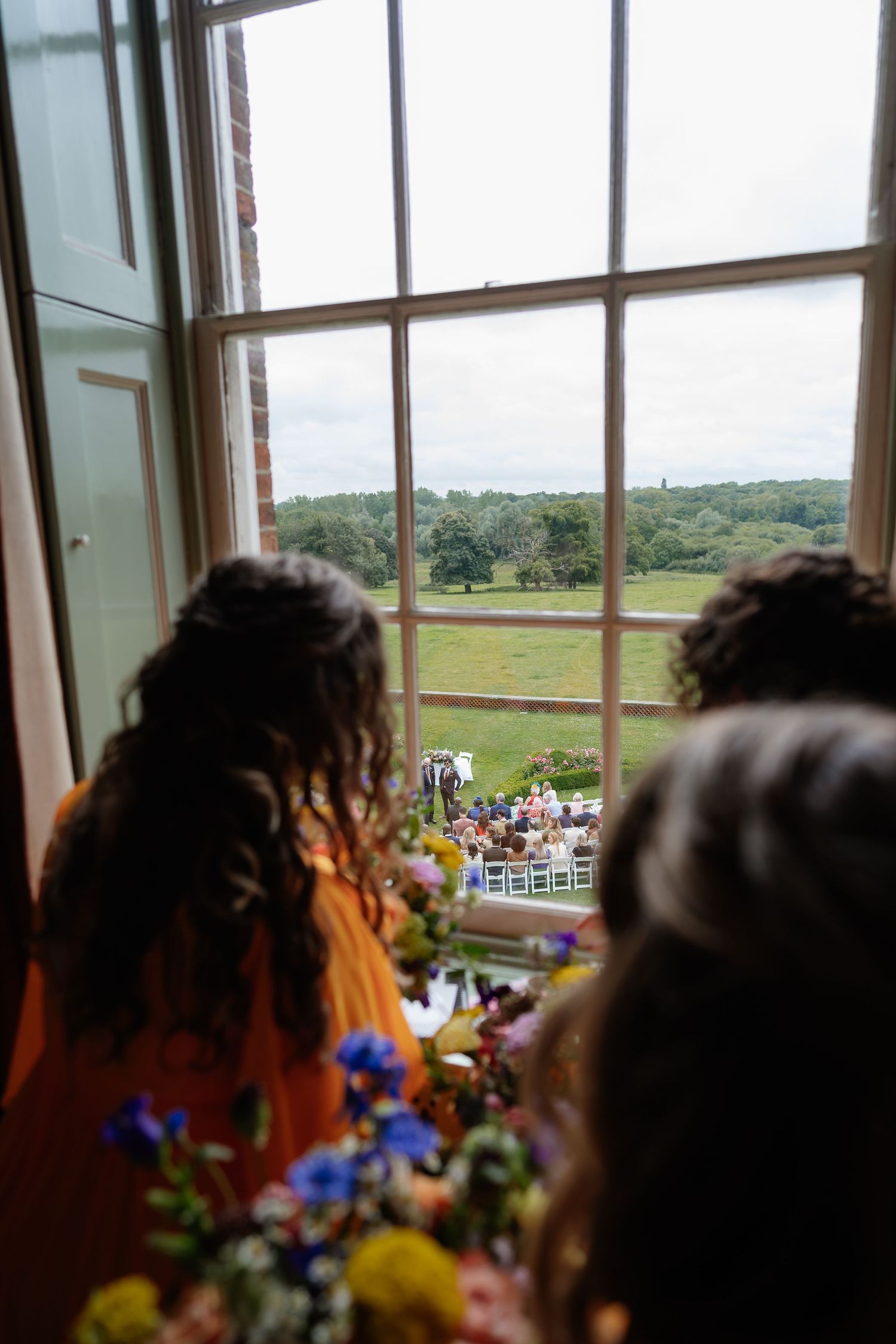 Bridesmaids in orange dresses watching outdoor wedding ceremony through large window overlooking garden.