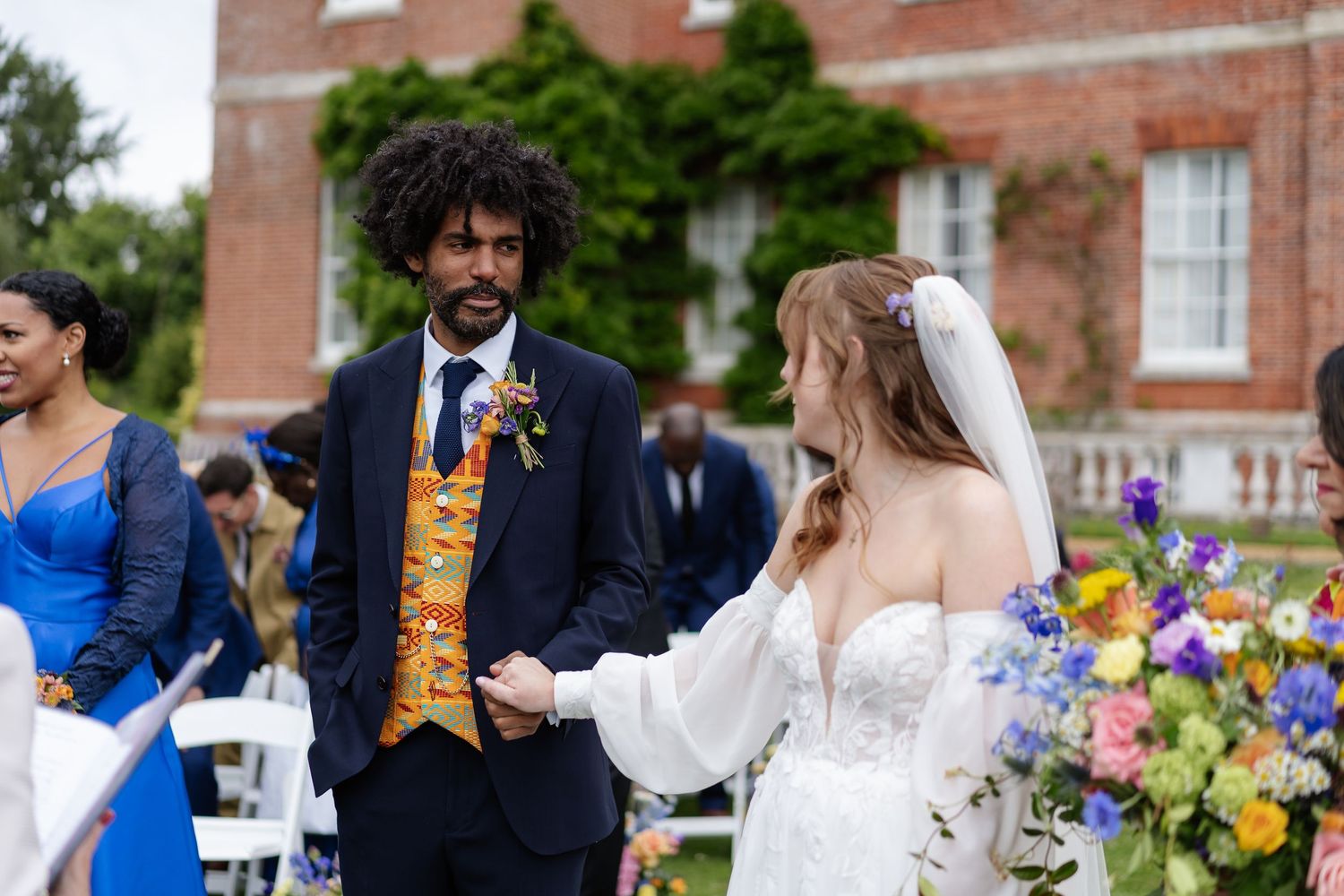 Bride and groom holding hands at outdoor wedding ceremony in front of elegant red brick manor house.