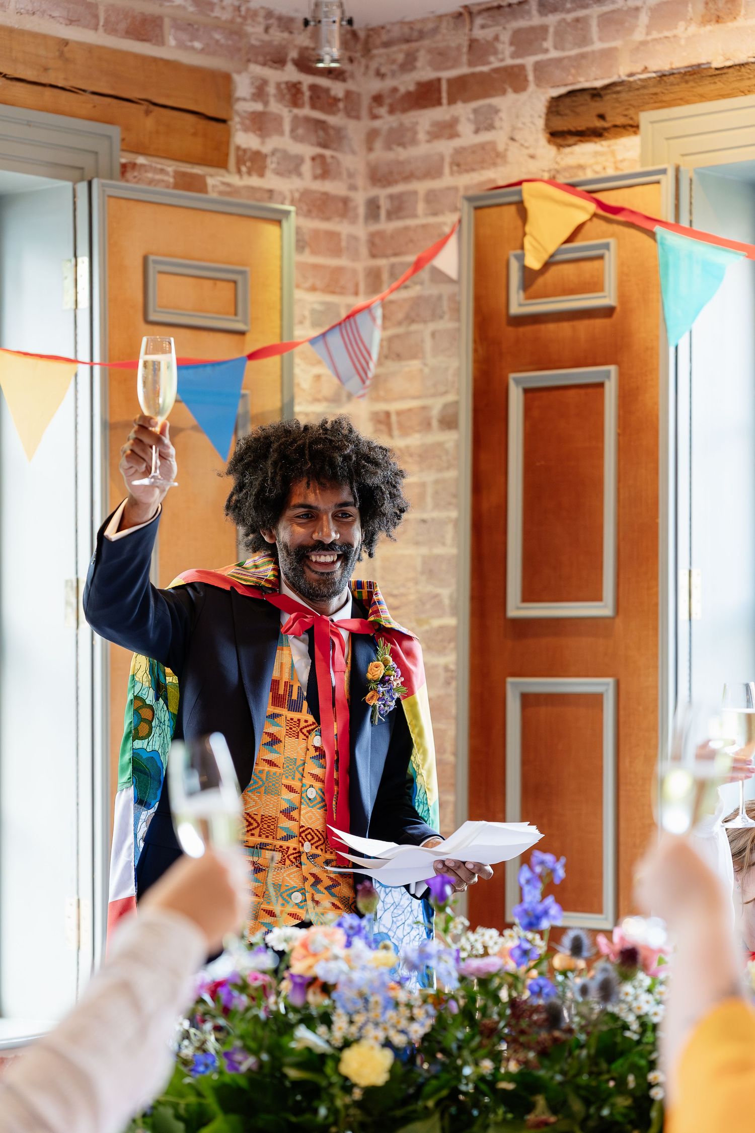 Groom in vibrant decorated jacket raises champagne glass during wedding toast surrounded by colorful bunting decorations.