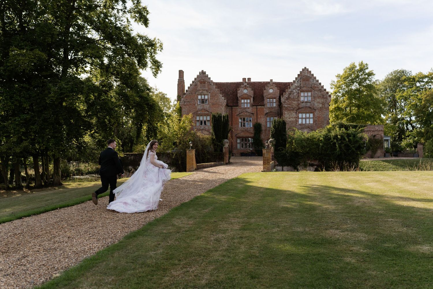 Bride and groom walk along gravel path toward grand red brick manor house on their wedding day.