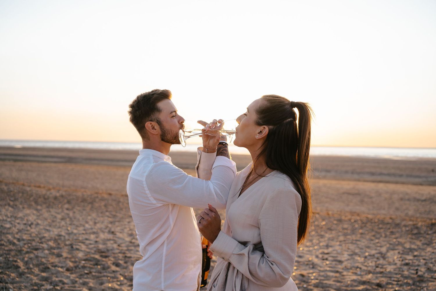 Couple toasting and drinking champagne face to face on a sunny beach at sunset.