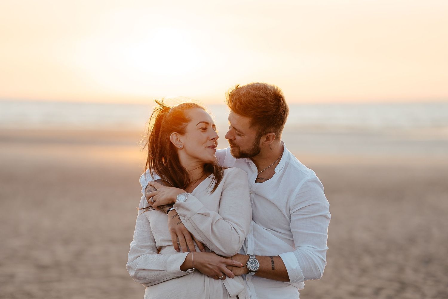 A couple sharing a tender romantic moment on a beach, embracing warmly during a glowing sunset.