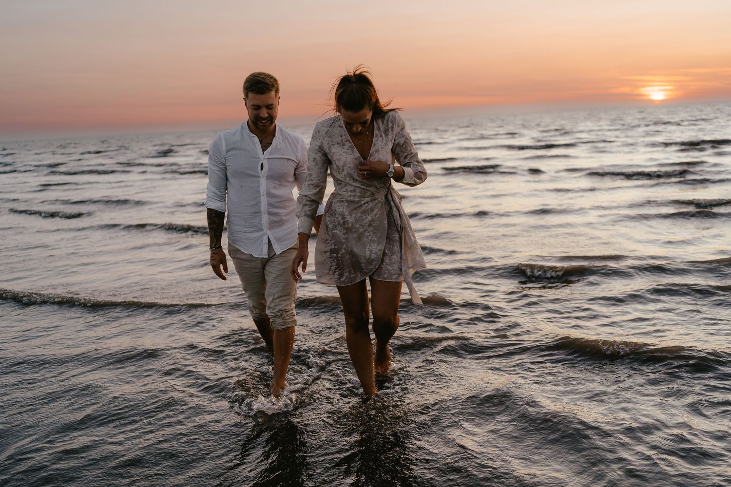 Couple walking hand in hand through shallow ocean waves at colorful sunset.