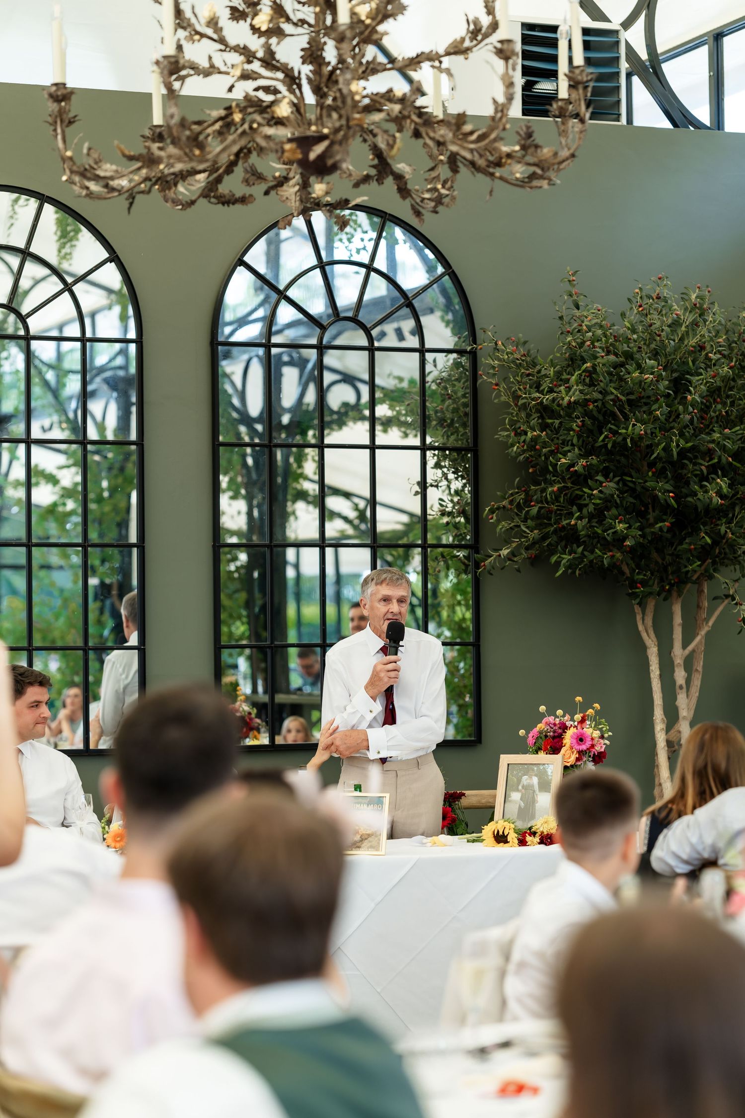 Man in white shirt gives a speech with microphone at elegant wedding reception with arched windows and chandelier.
