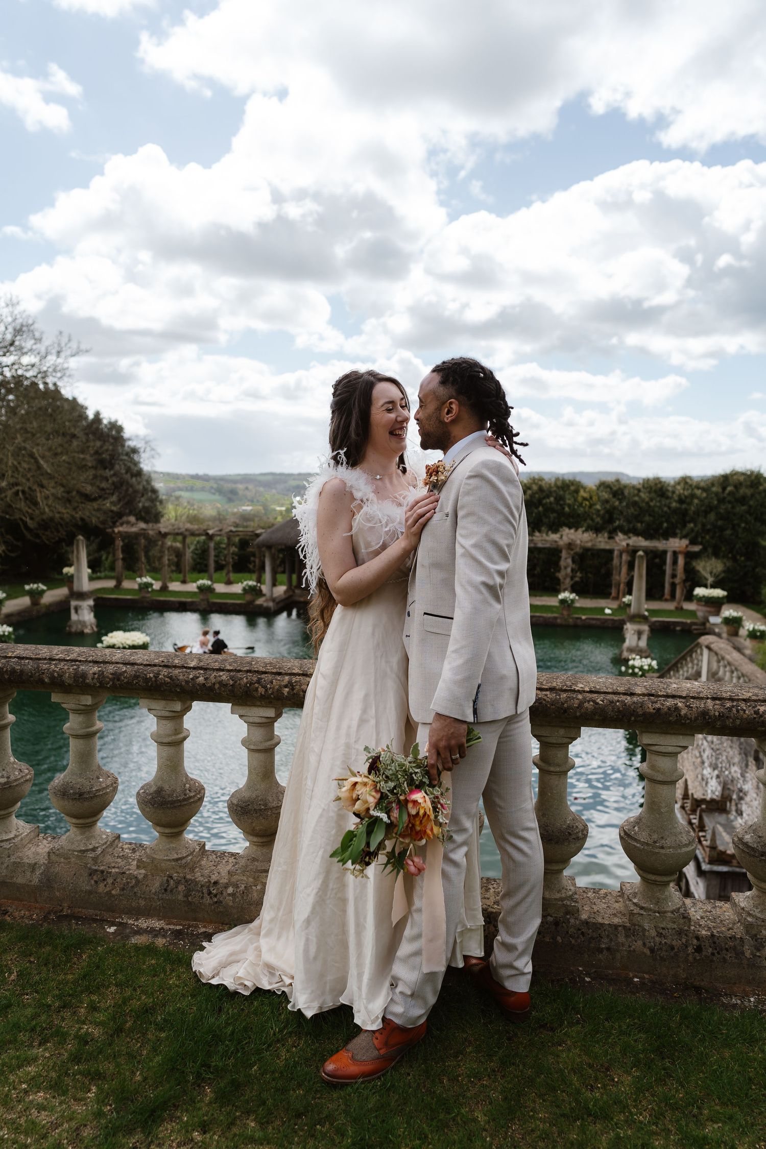 Bride and groom sharing a romantic moment on a stone balustrade overlooking scenic countryside gardens.