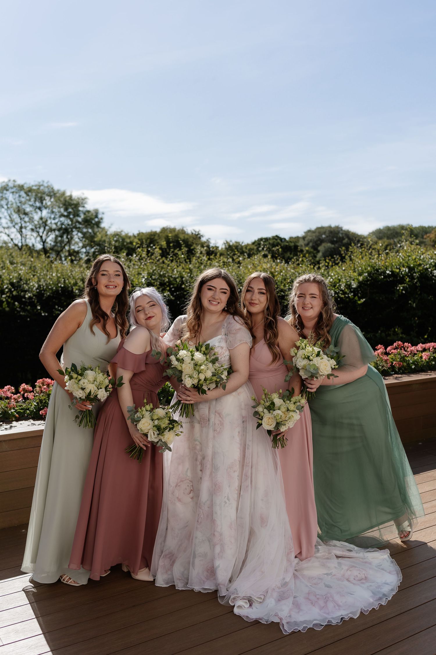 Bride and four bridesmaids in pink and green dresses holding white bouquets outdoors.