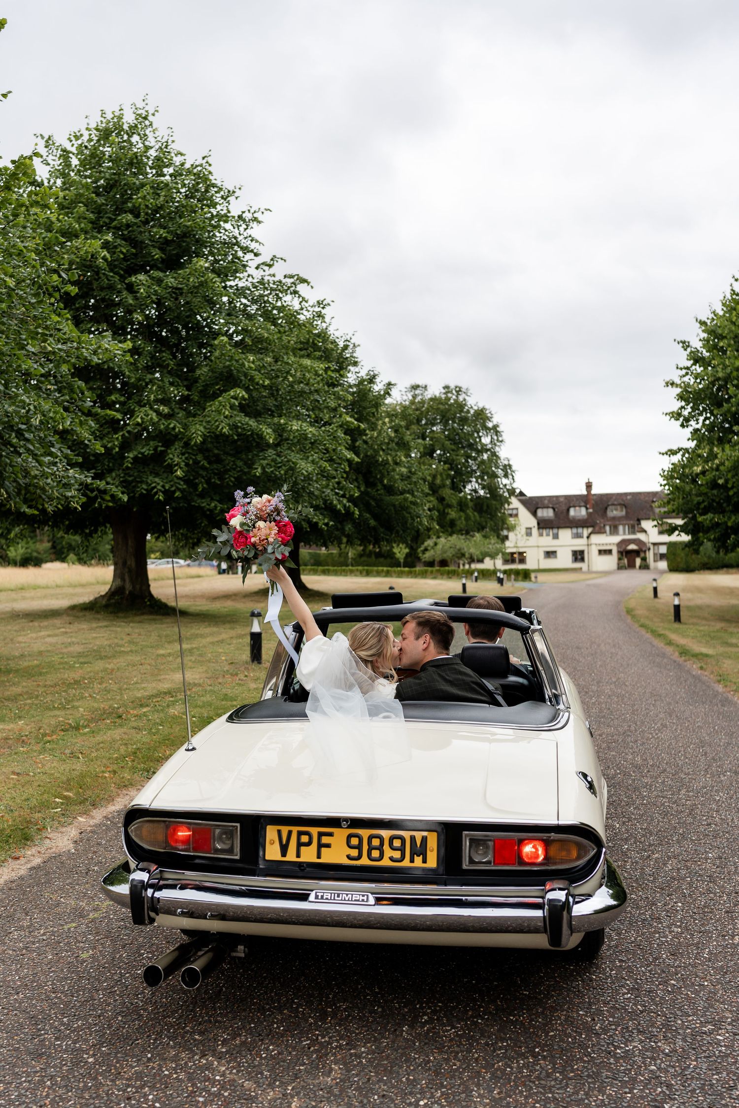 Newlyweds kiss in vintage white convertible car raising bouquet as they drive from wedding venue.