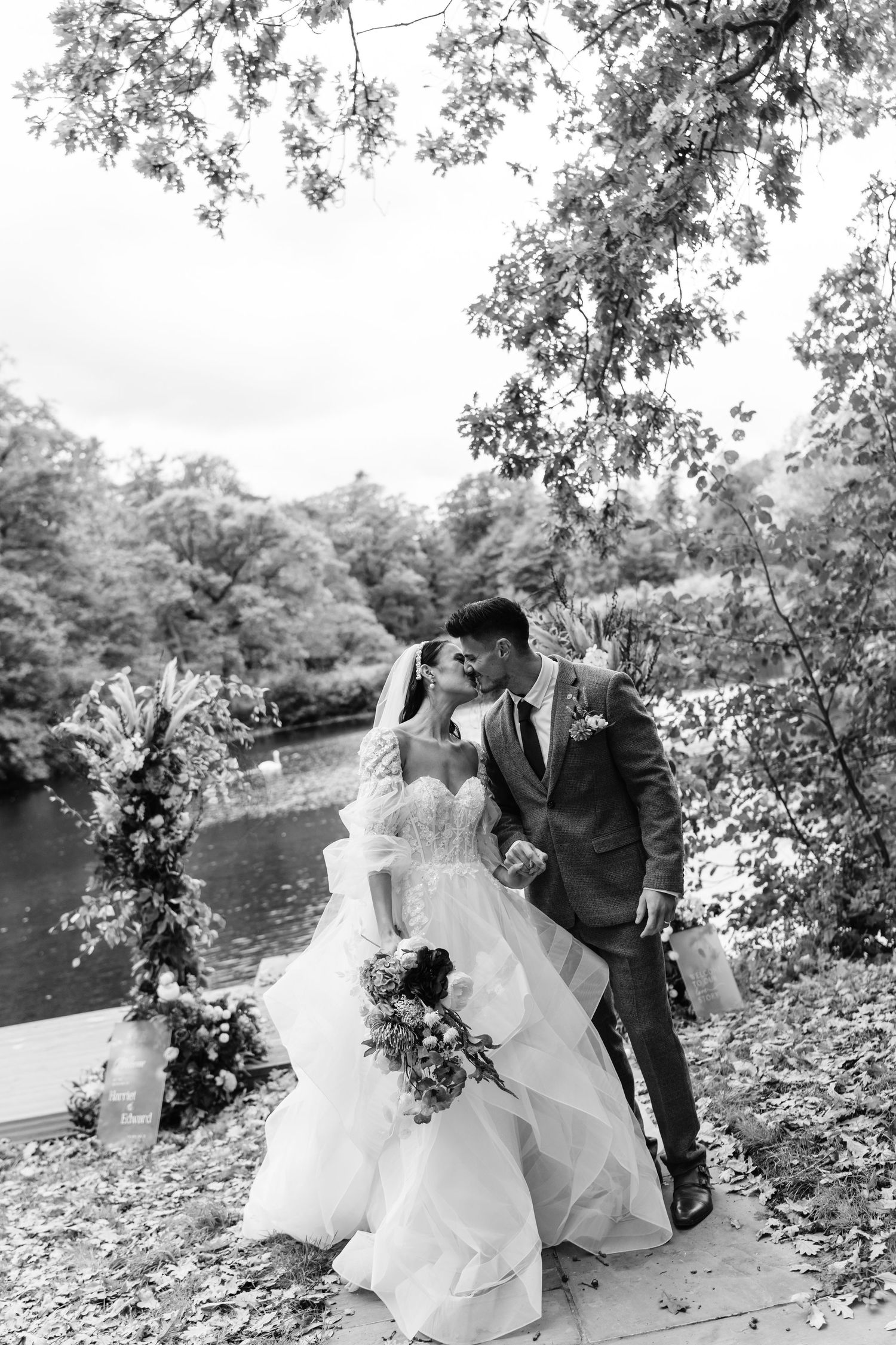 Black and white photo of a bride and groom kissing at their outdoor wedding ceremony by a lake.
