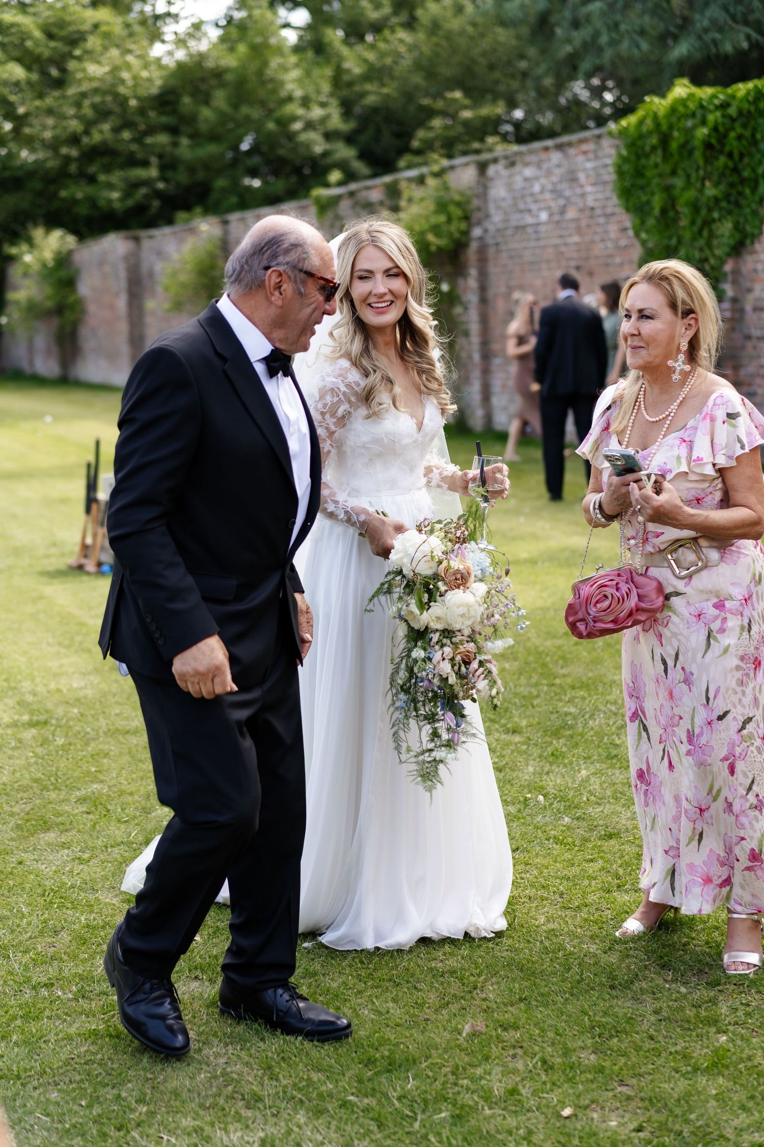 Smiling bride in lace wedding gown holding cascading bouquet chatting with guests on garden lawn.